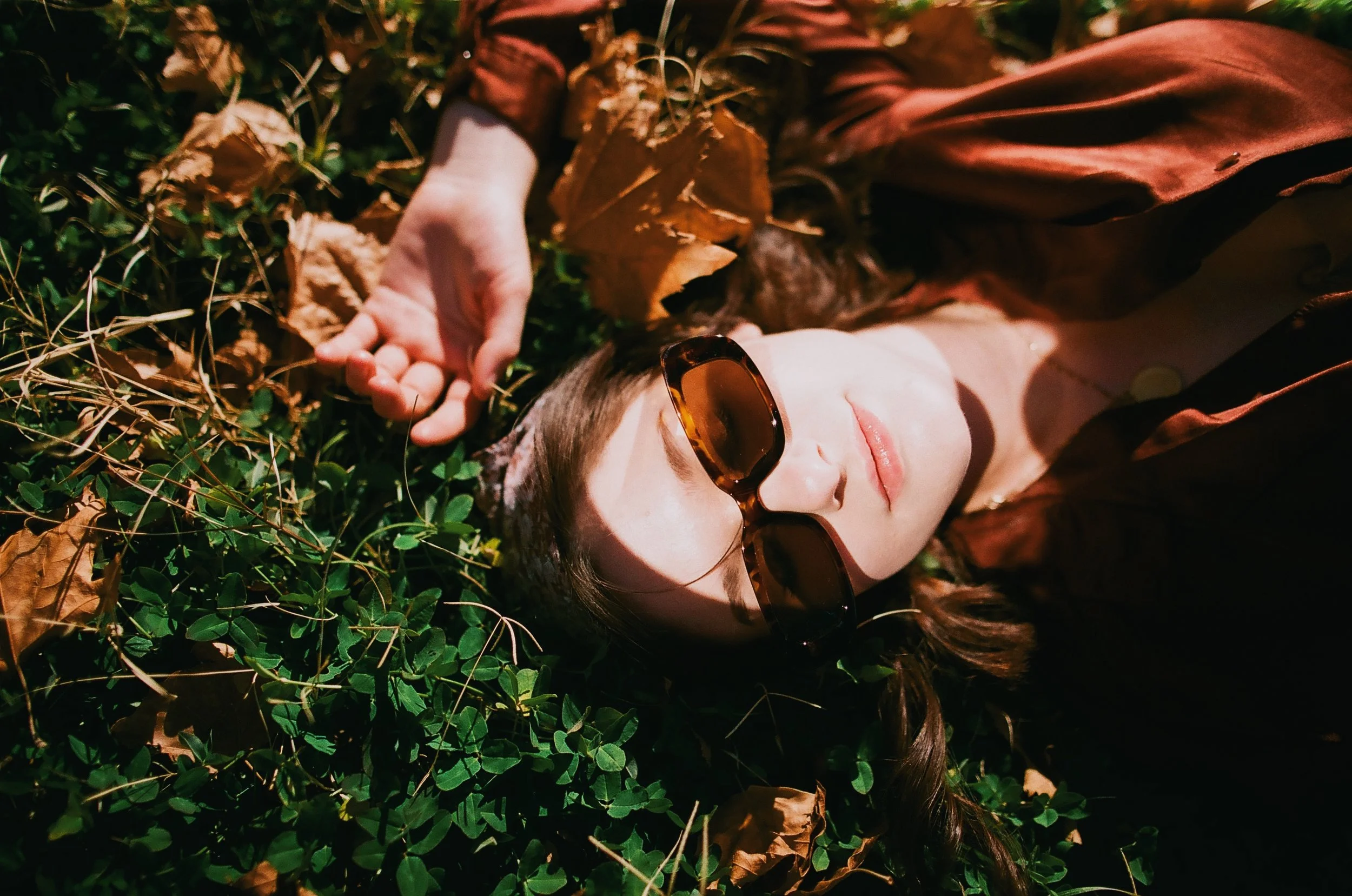 A woman with brown hair and sunglasses lying on the grass among dry leaves and green plants.