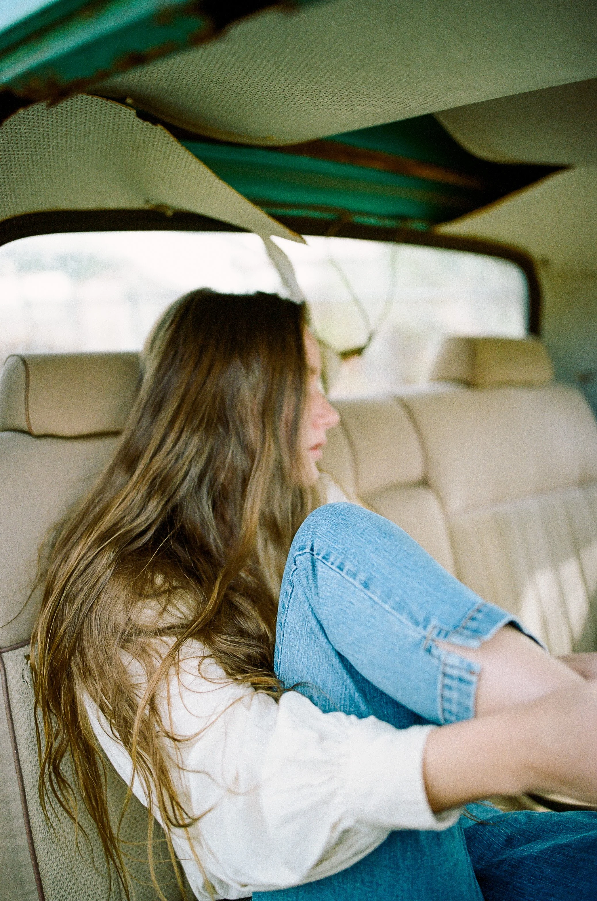 A woman with long, wavy hair sitting in the backseat of a vehicle, looking out the window, wearing a white long-sleeve shirt and blue jeans.