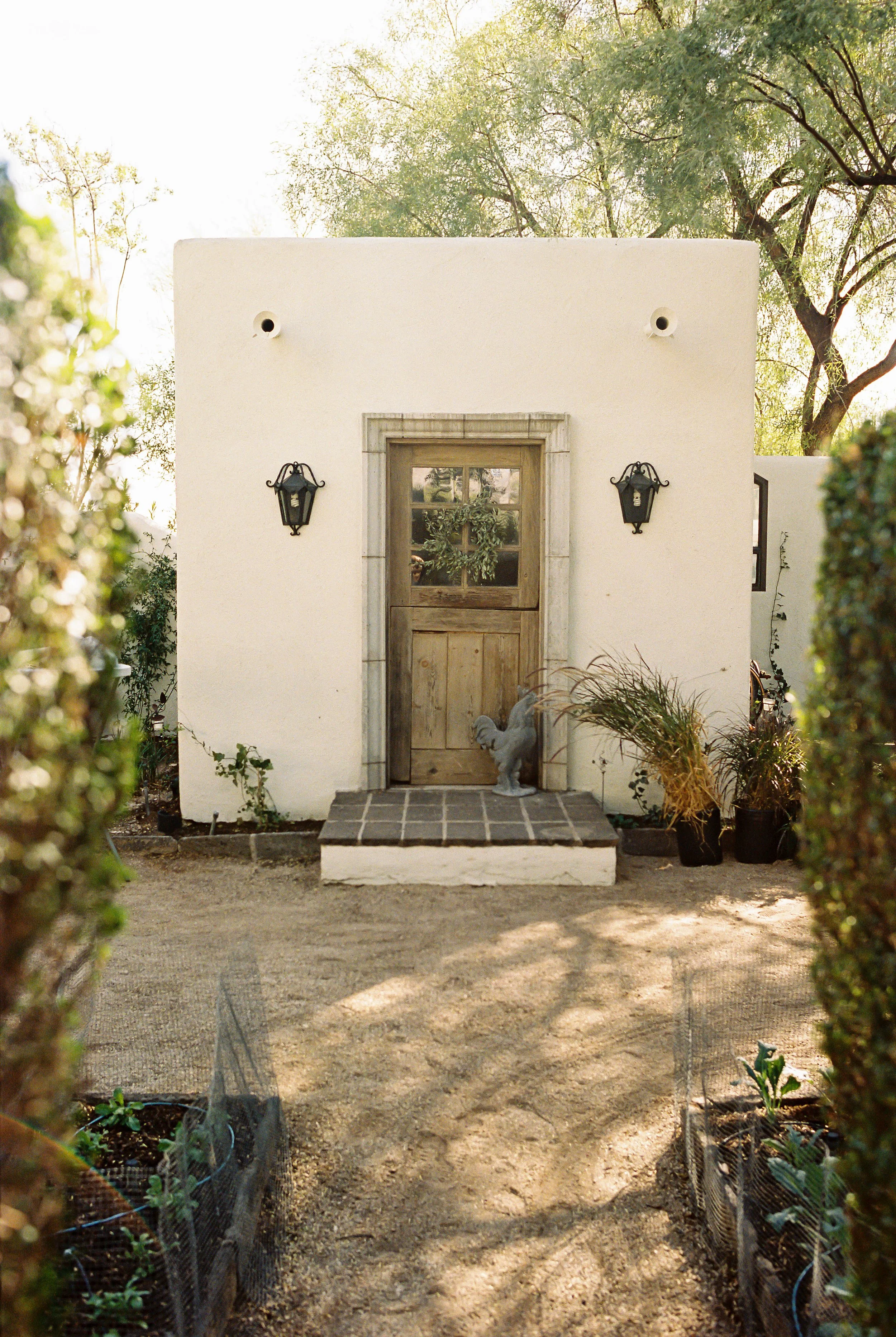 Front view of a small white stucco house with a wooden door, two black lanterns on either side, potted plants, and a rooster statue on the porch, surrounded by greenery and trees.