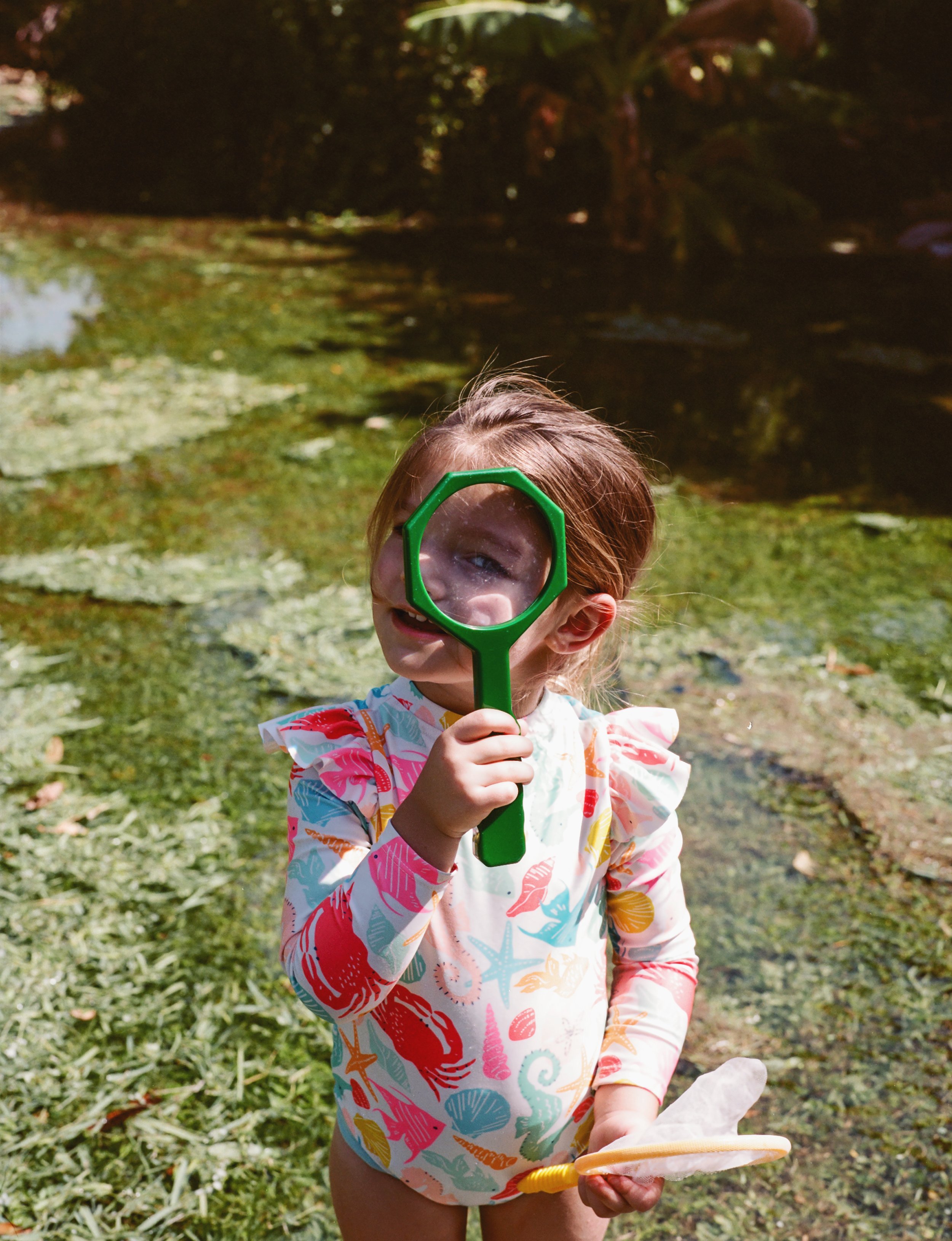 Child playing outdoors, holding a green magnifying glass and a yellow toy shovel, wearing a colorful long-sleeve shirt with sea creatures pattern, in front of a pond with lily pads.