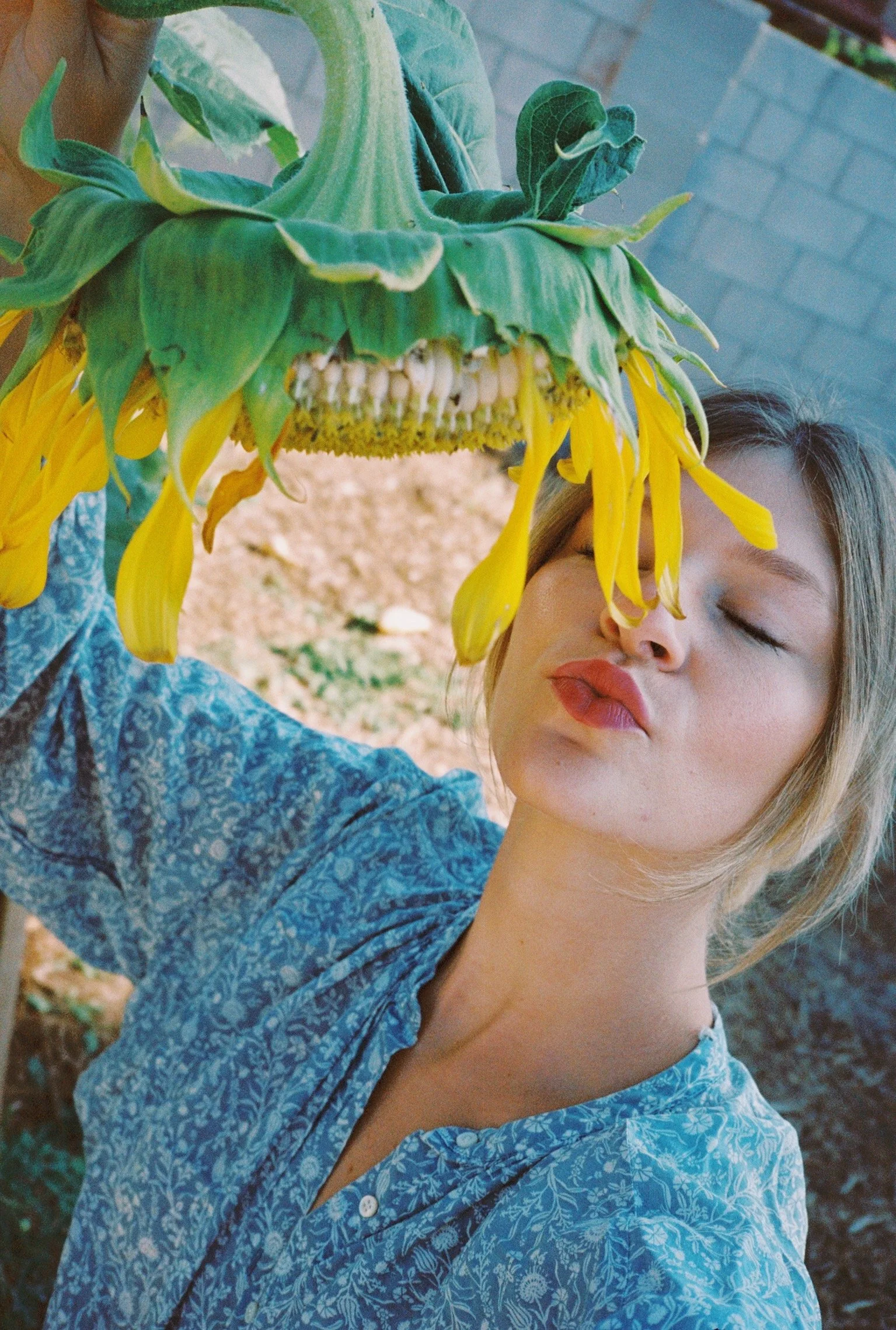 A young woman with pale skin and blonde hair is making a kissing face with her eyes closed, holding a large, hanging sunflower upside down close to her face.