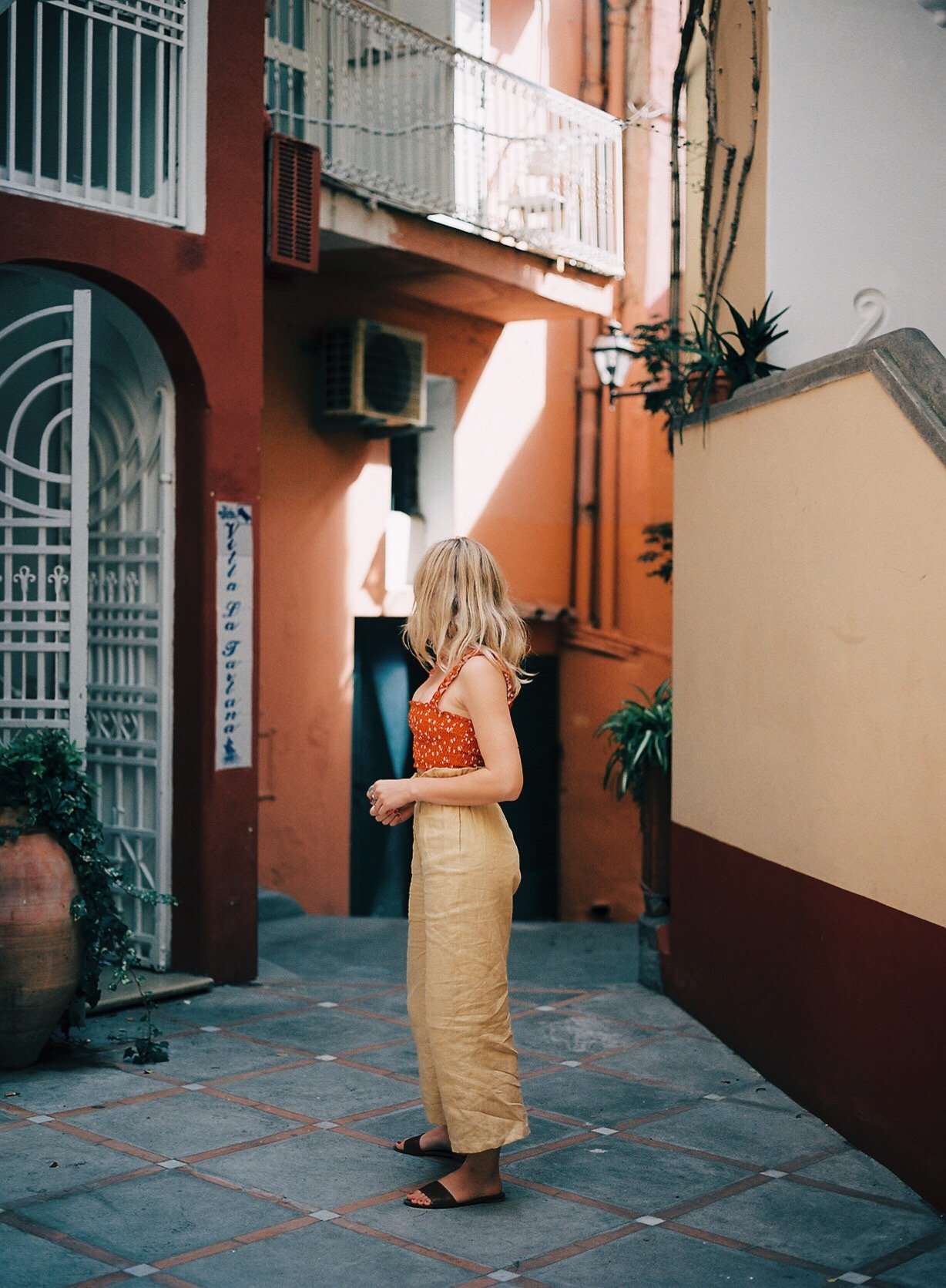 A woman with blonde hair standing in an outdoor alleyway with vibrant colored walls and potted plants.