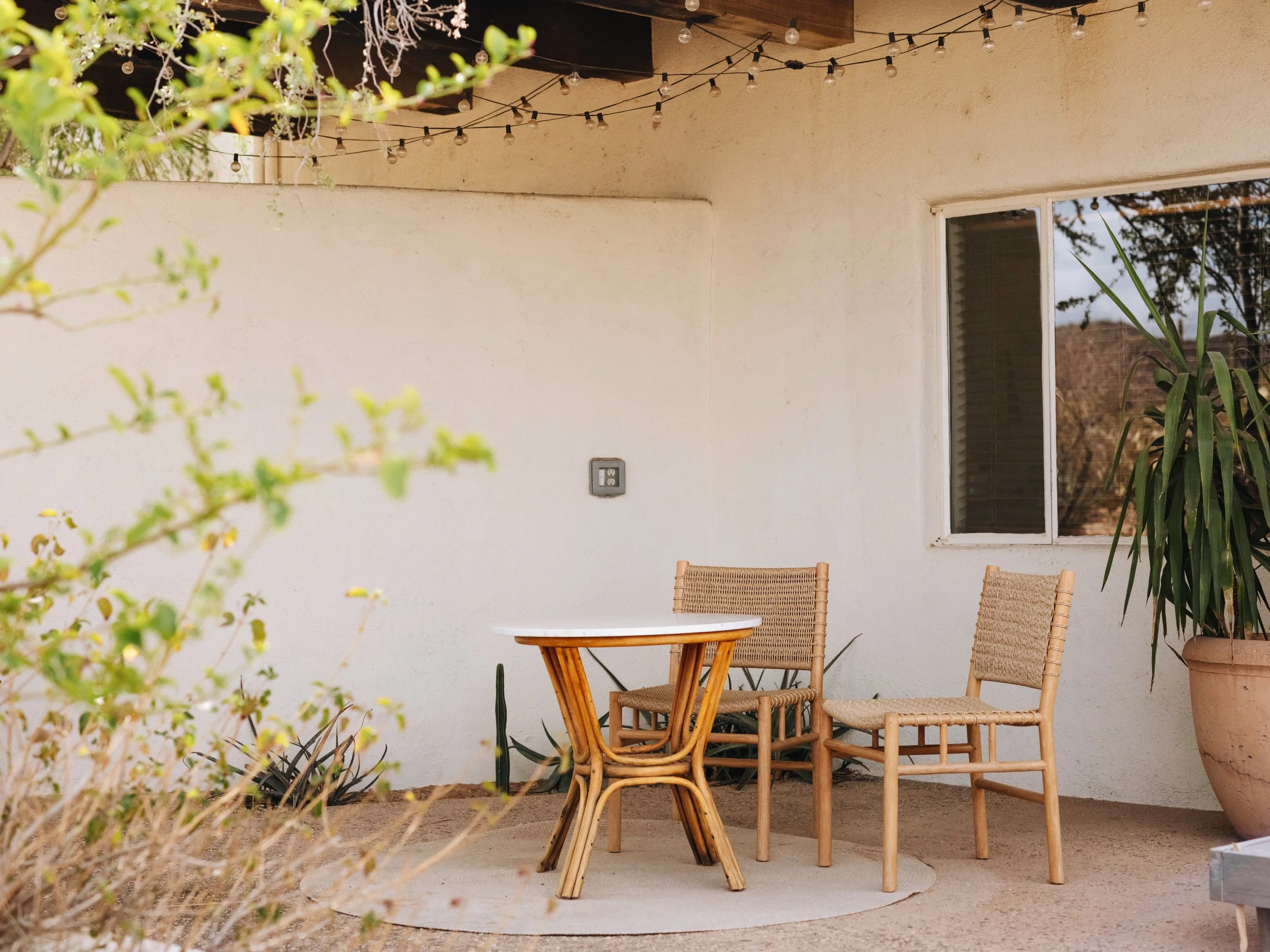 Outdoor patio with a round white table and three wicker chairs, large potted plant, and string lights hanging above, against a beige wall with a window.