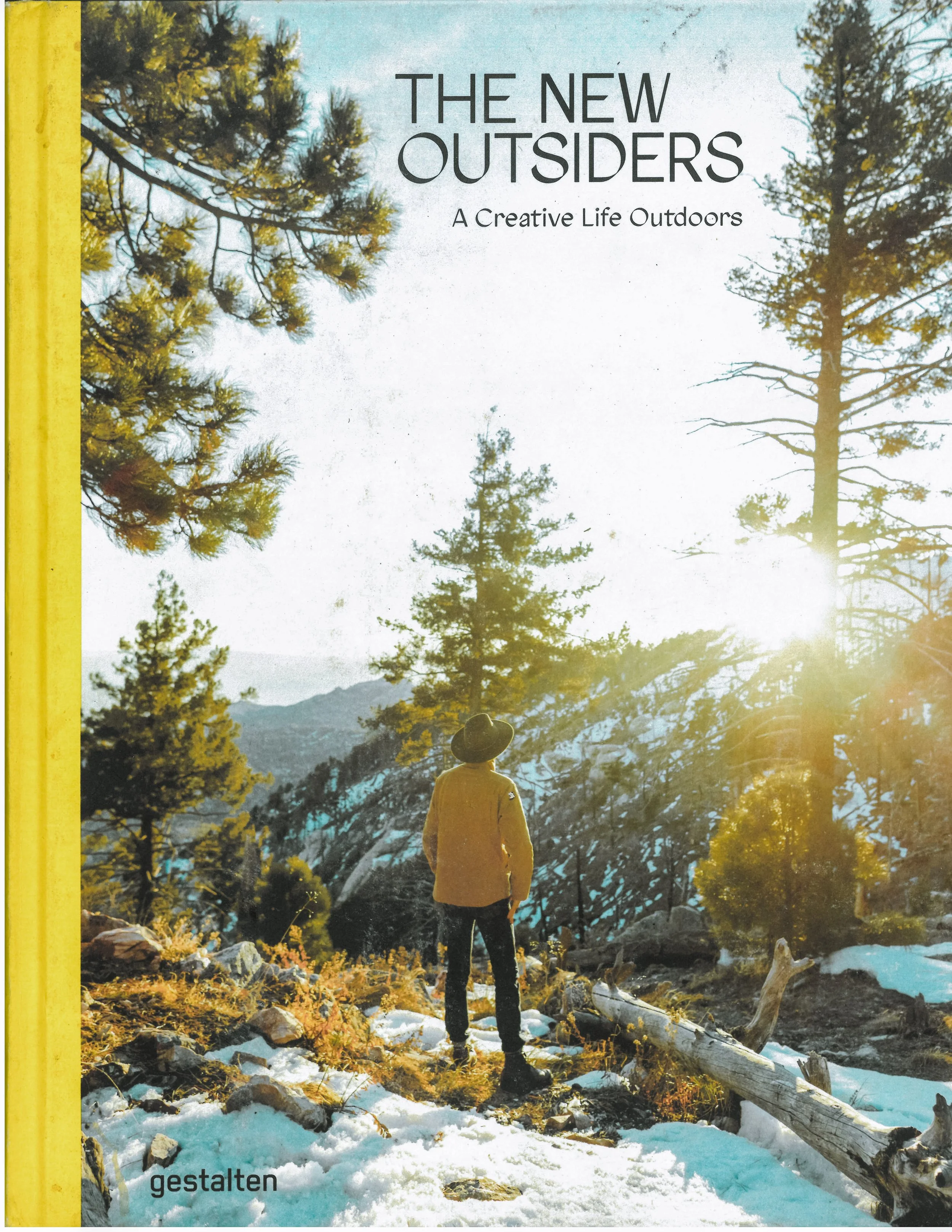 Book cover titled 'The New Outsiders: A Creative Life Outdoors' showing a person in a yellow jacket and wide-brimmed hat standing in a snow-covered forest with tall pine trees and mountains in the background, backlit by sunlight.