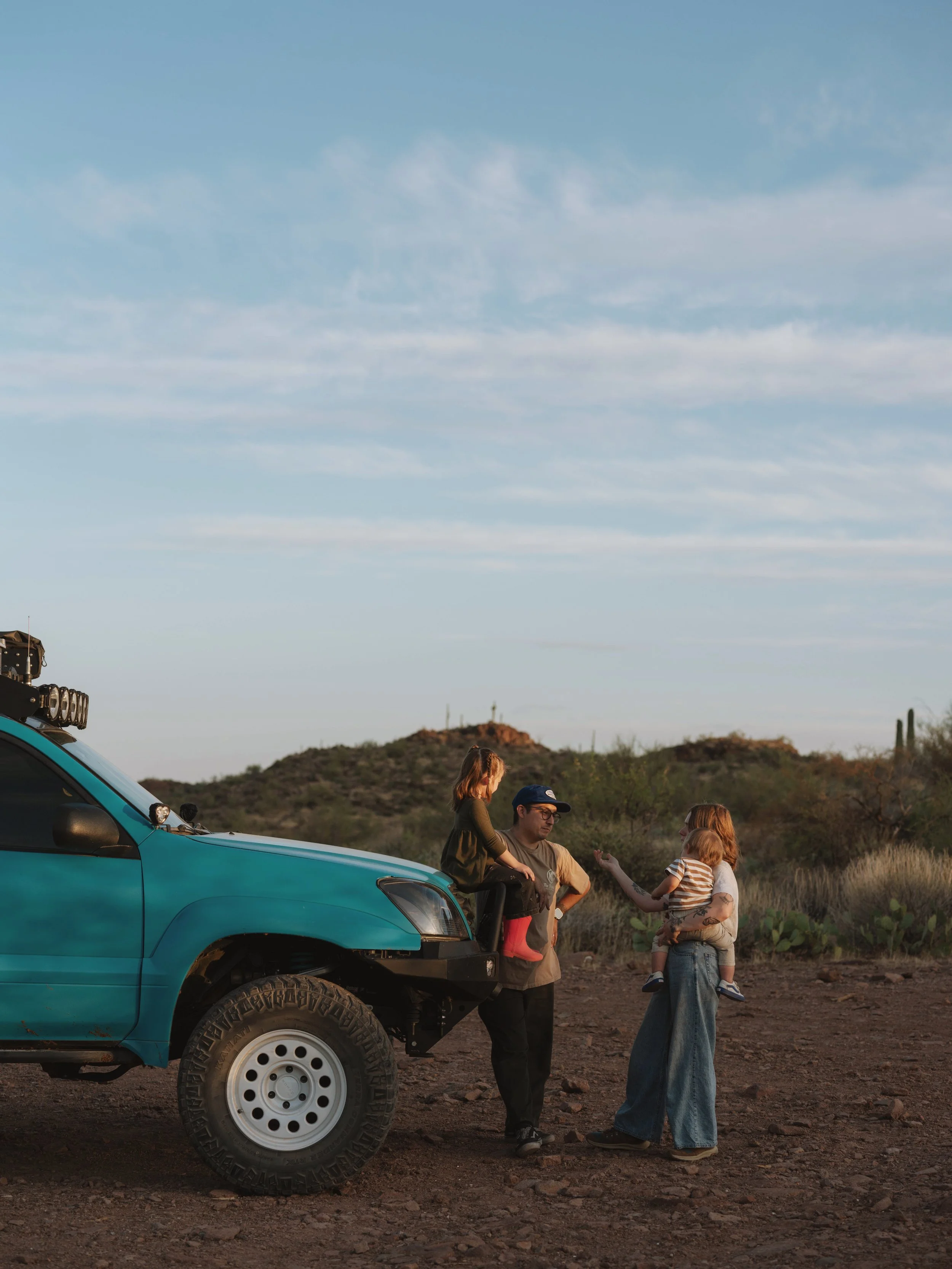 A family of four, including two children, standing next to an off-road vehicle in a desert landscape during sunset.