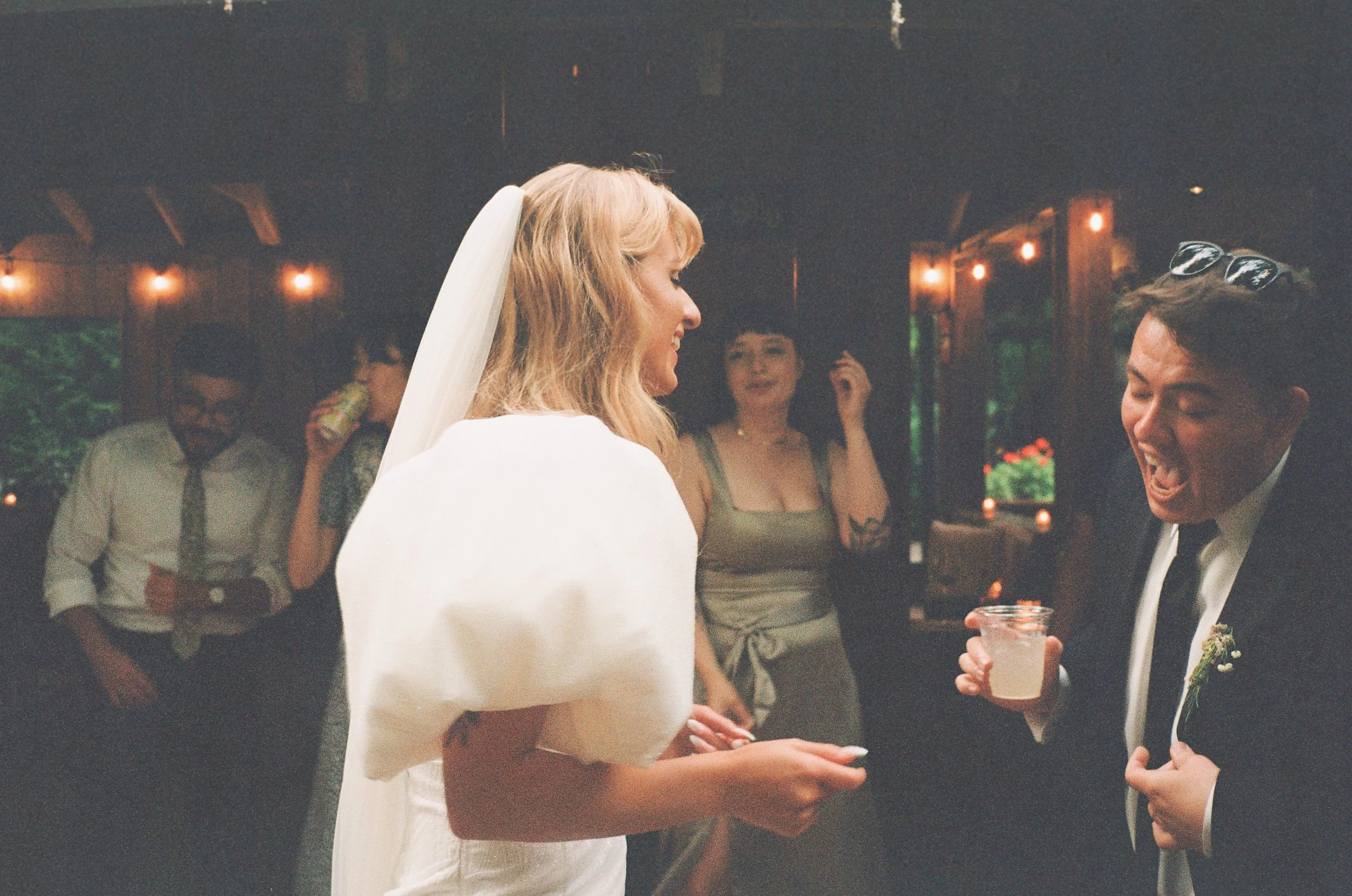 A bride in a white wedding gown and veil laughing and talking to a groom in a tuxedo at a wedding reception, with guests dancing and enjoying drinks in the background.