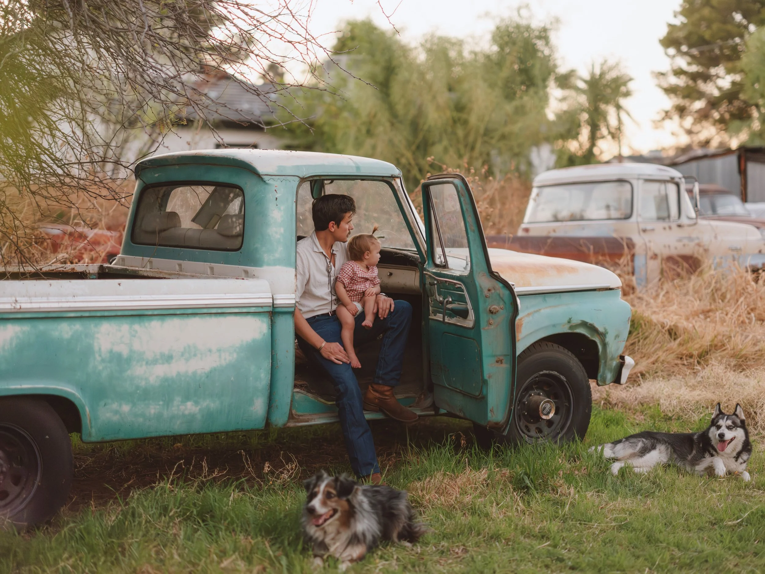 A man and a young child sitting inside an old, weathered blue pickup truck with its door open, parked in a grassy area. Two dogs are lying on the grass nearby, and there are additional old vehicles in the background, along with trees and a house.