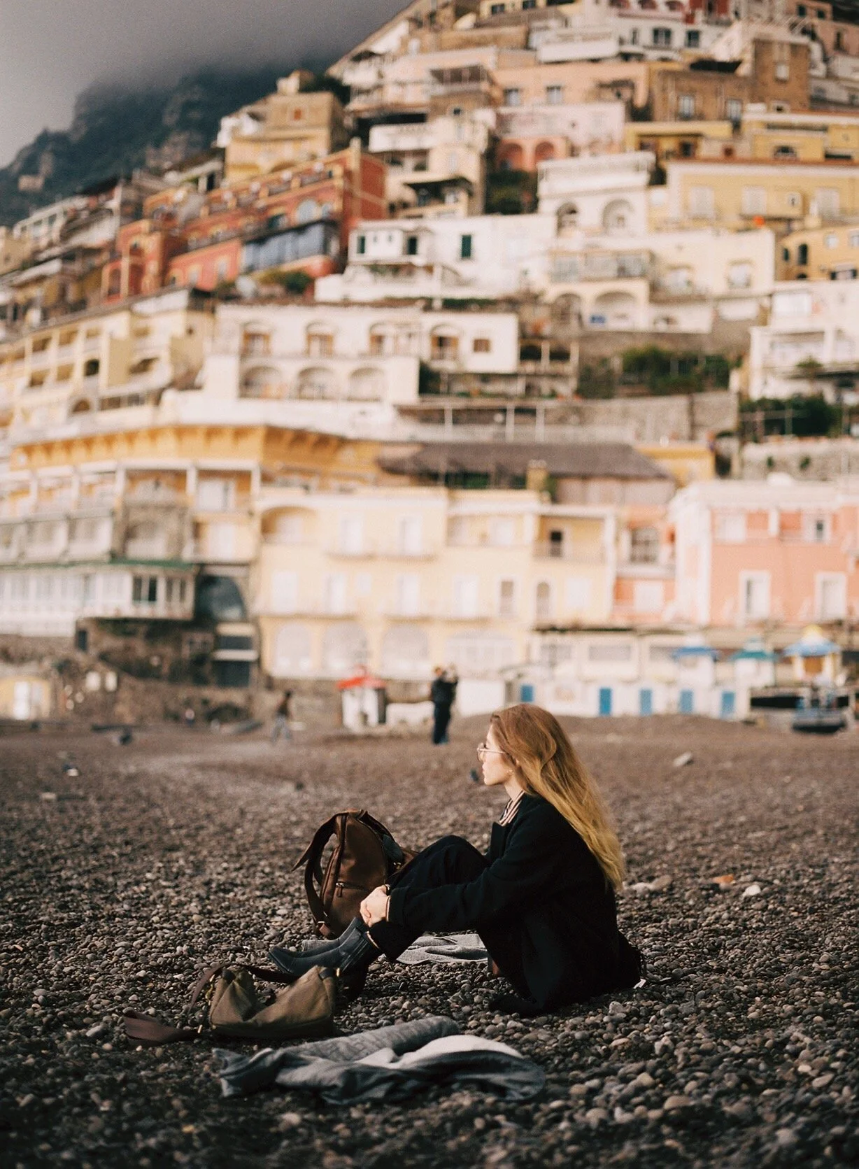 A young woman with long hair and glasses sitting on a pebble beach, packing her backpack with colorful houses built into a hillside in the background.