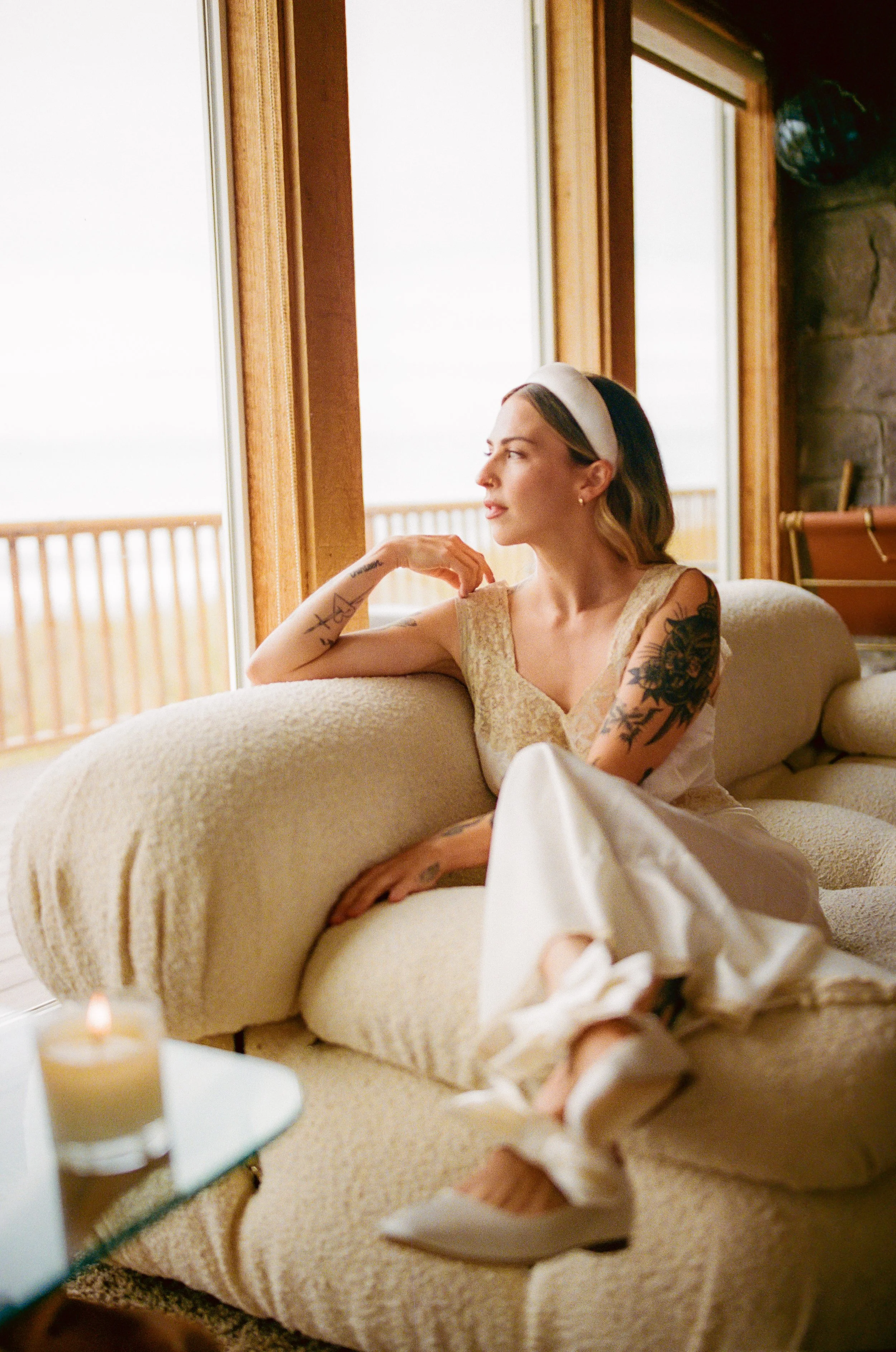 A woman with styled hair, tattoos, and a headband sitting on a beige couch by a large window, gazing outside with a serene expression.