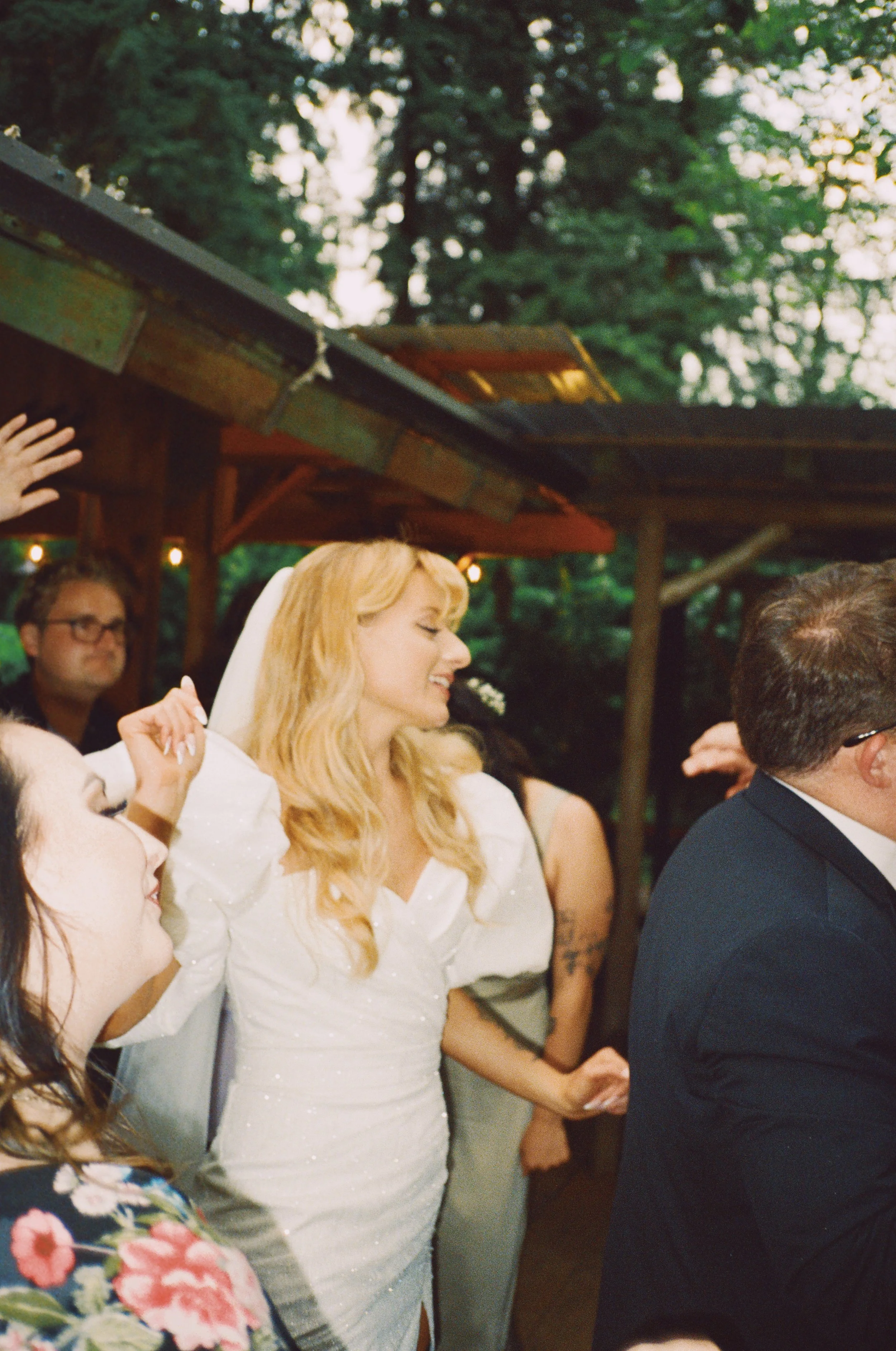 A bride with blonde hair in a white dress and veil smiling and dancing at an outdoor wedding reception.