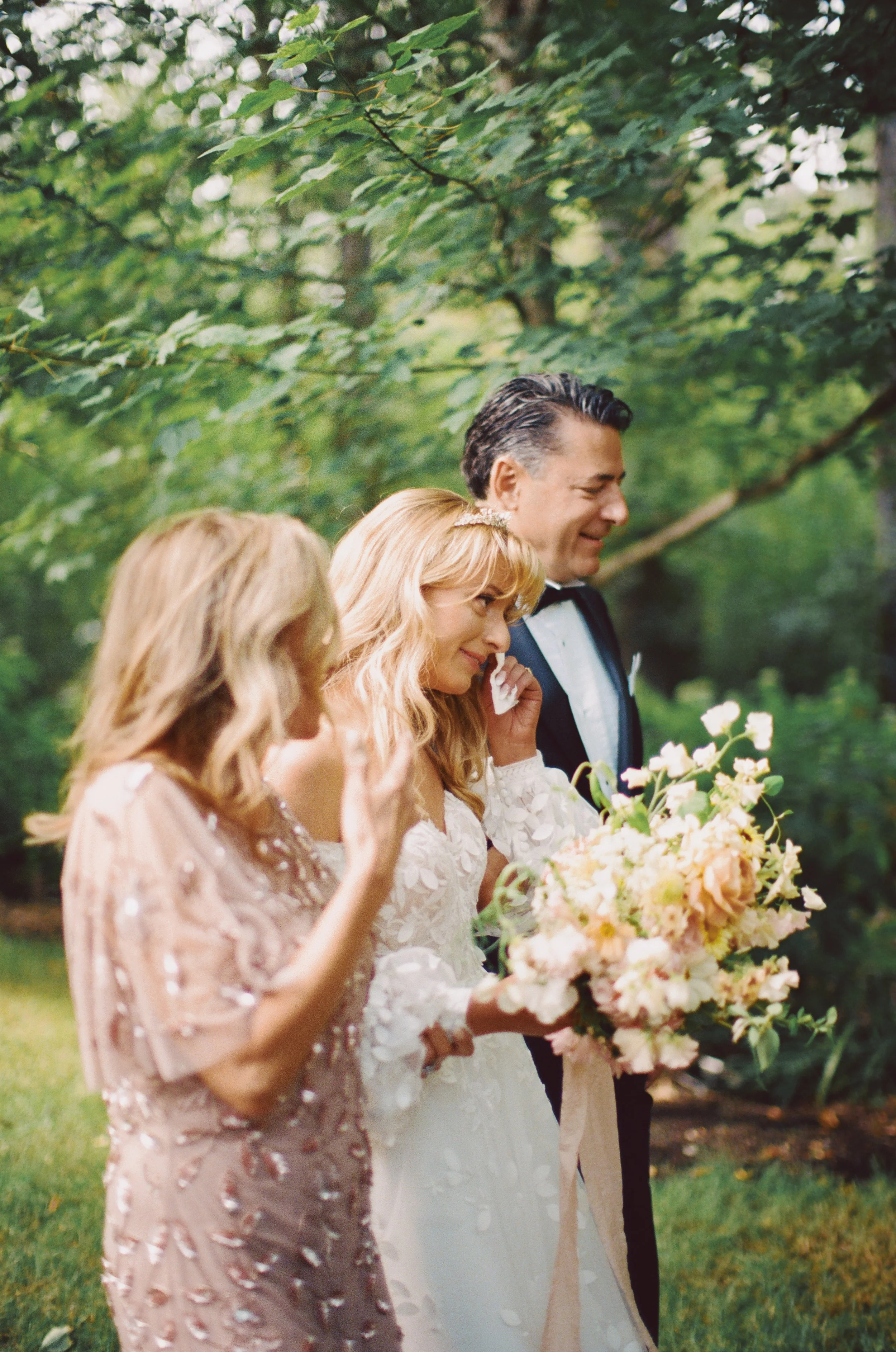 A wedding scene with a bride in a white dress holding a bouquet of light pink and peach flowers, a man in a tuxedo smiling, and a woman in a pink dress with embroidery, in a lush green outdoor setting.