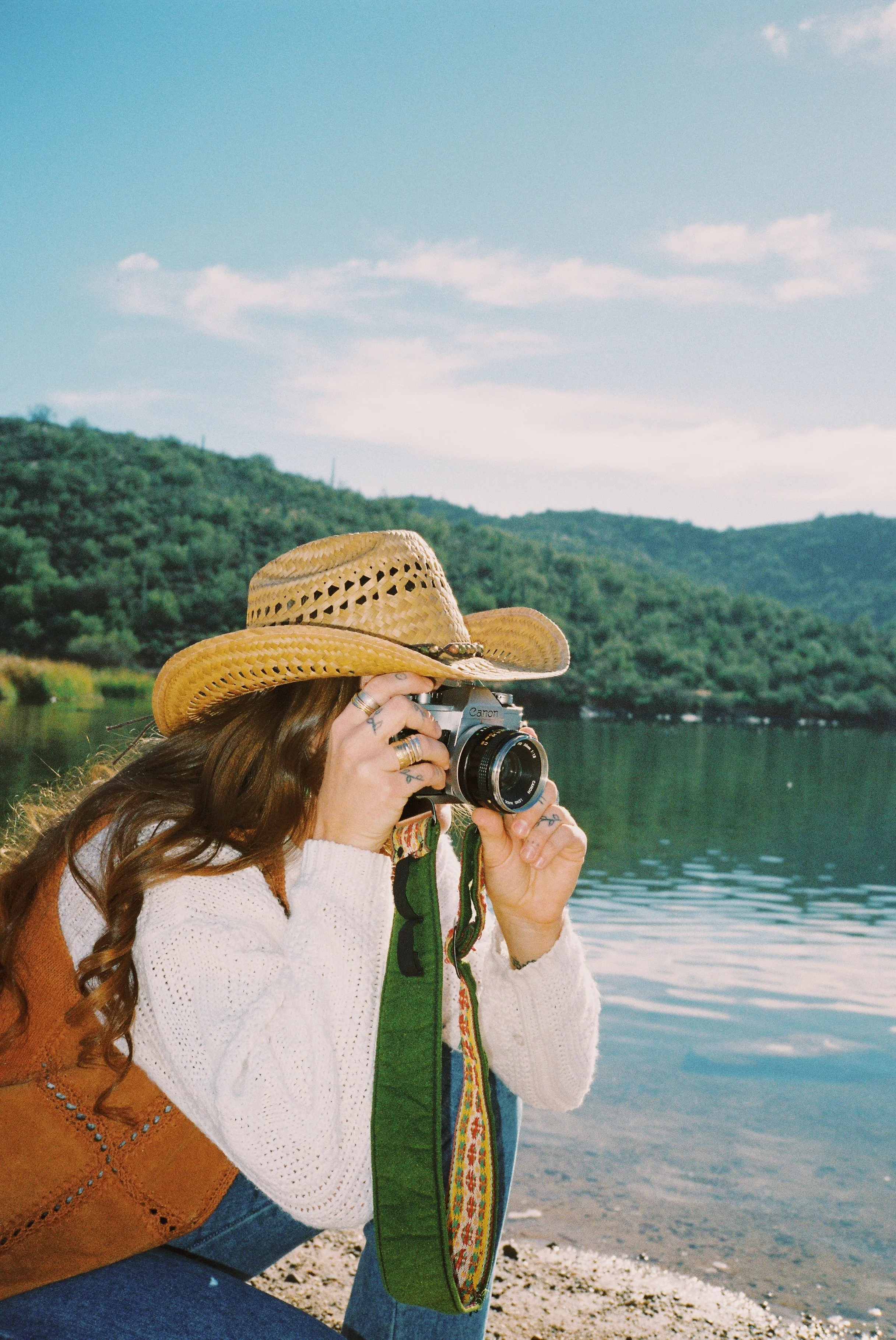 Woman wearing a straw hat taking a photograph with a camera near a lake, with green hills and blue sky in the background.
