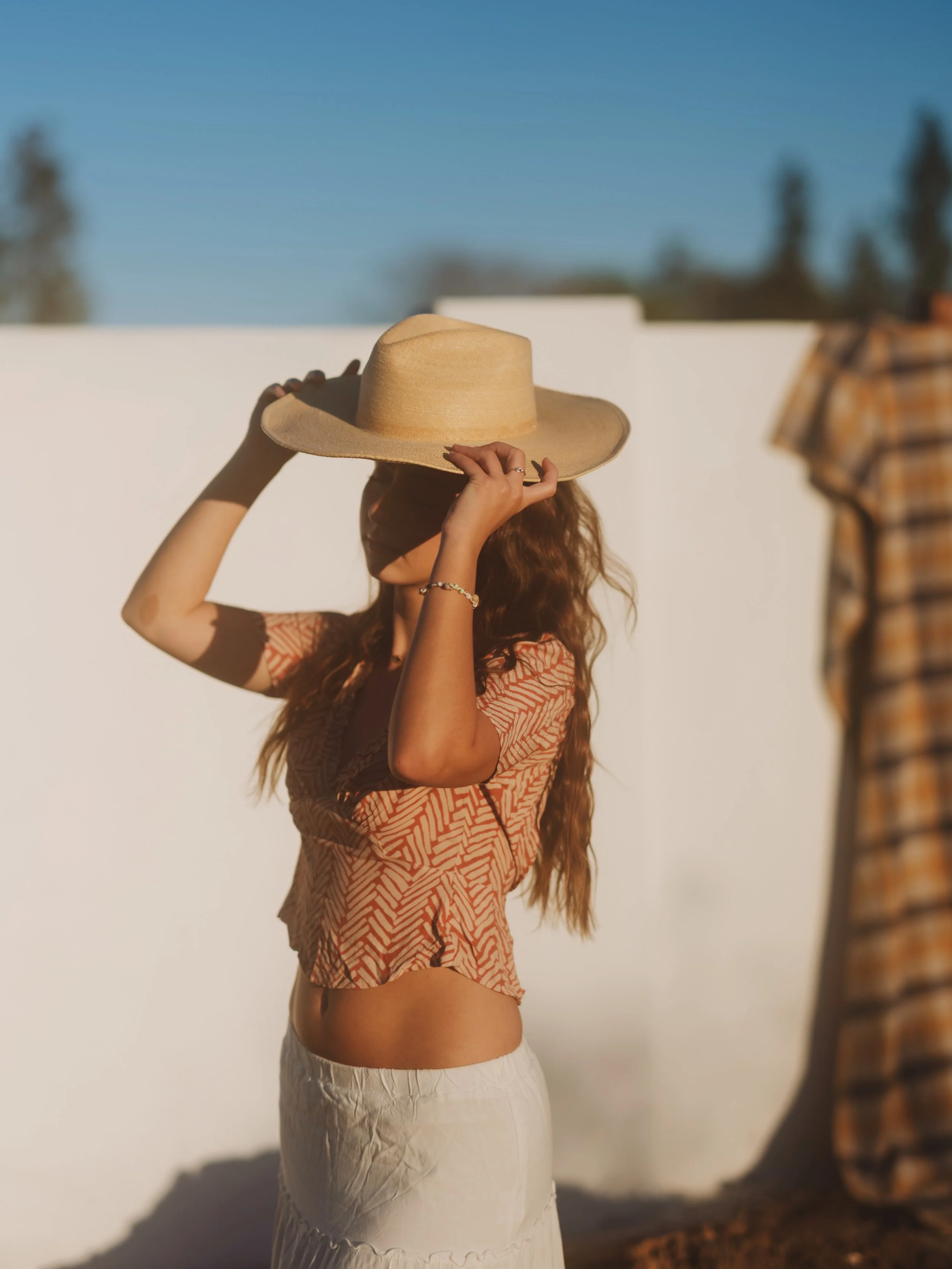 A woman wearing a hat adjusts it while standing outdoors in sunlight, wearing a patterned crop top and white skirt, with a white wall and trees in the background.