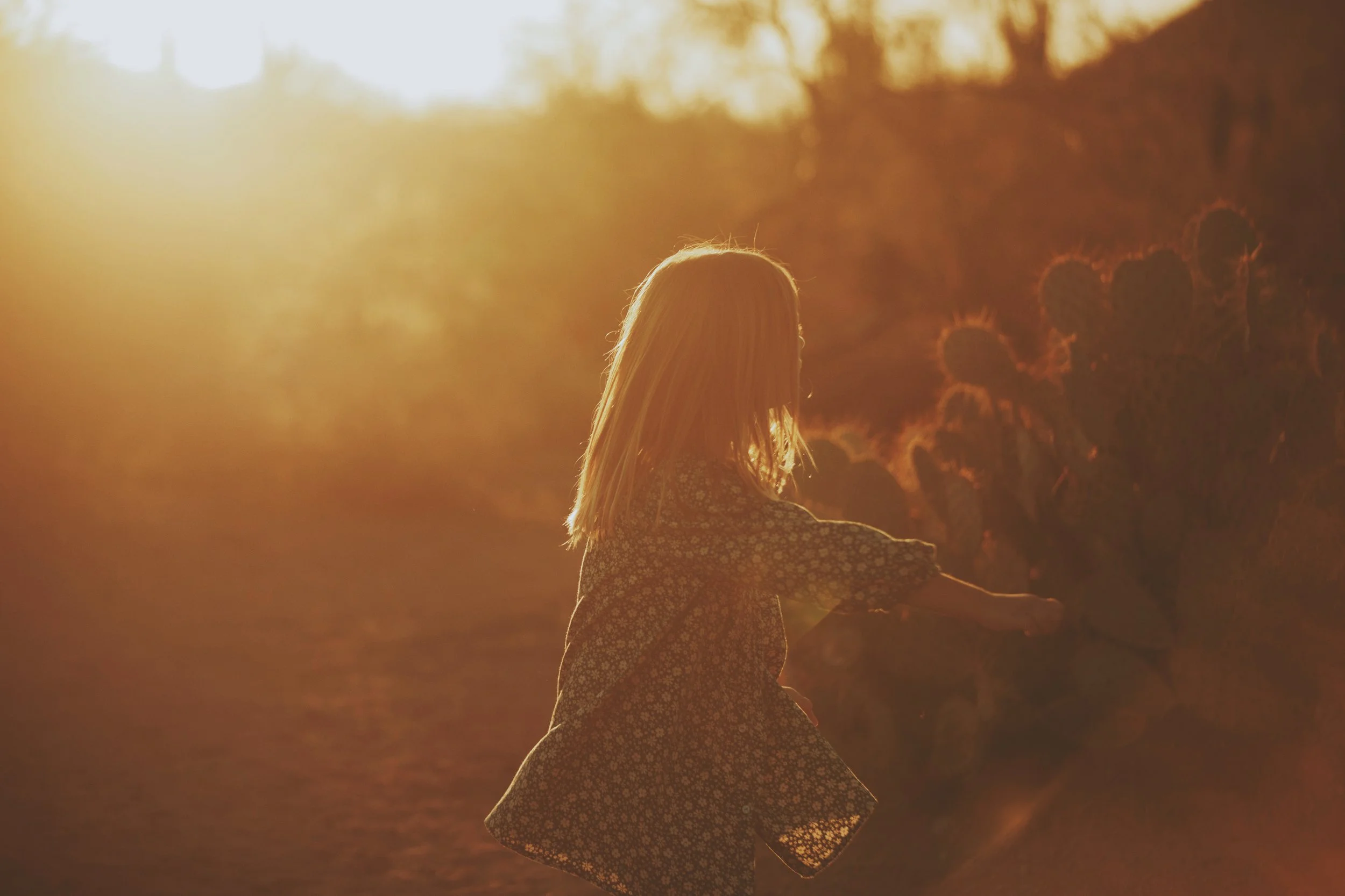 A young girl with long blonde hair playing outdoors at sunset, surrounded by cacti.