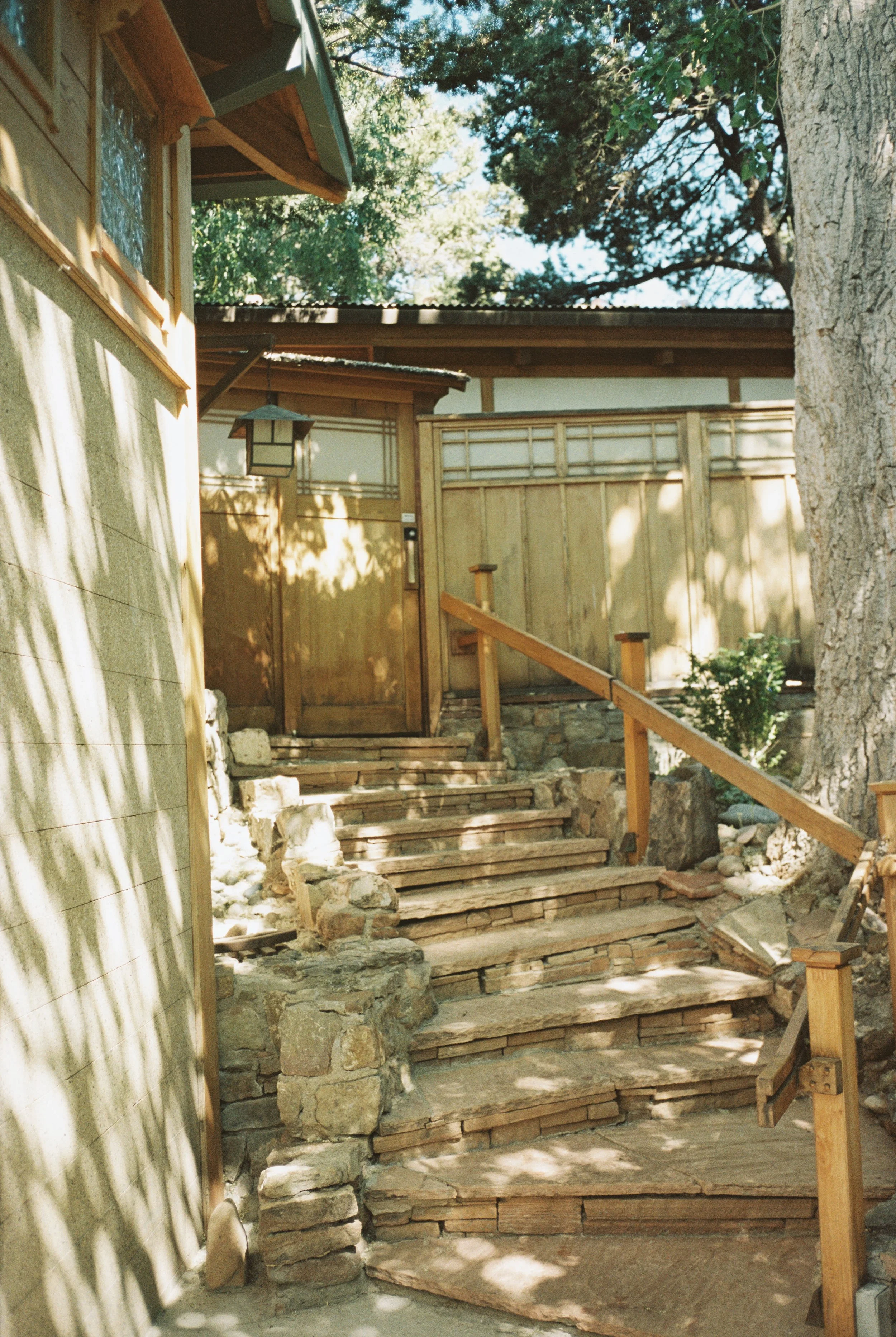 Wooden stairs leading to a door in a house, surrounded by a wooden fence and trees with shade and sunlight patterns.