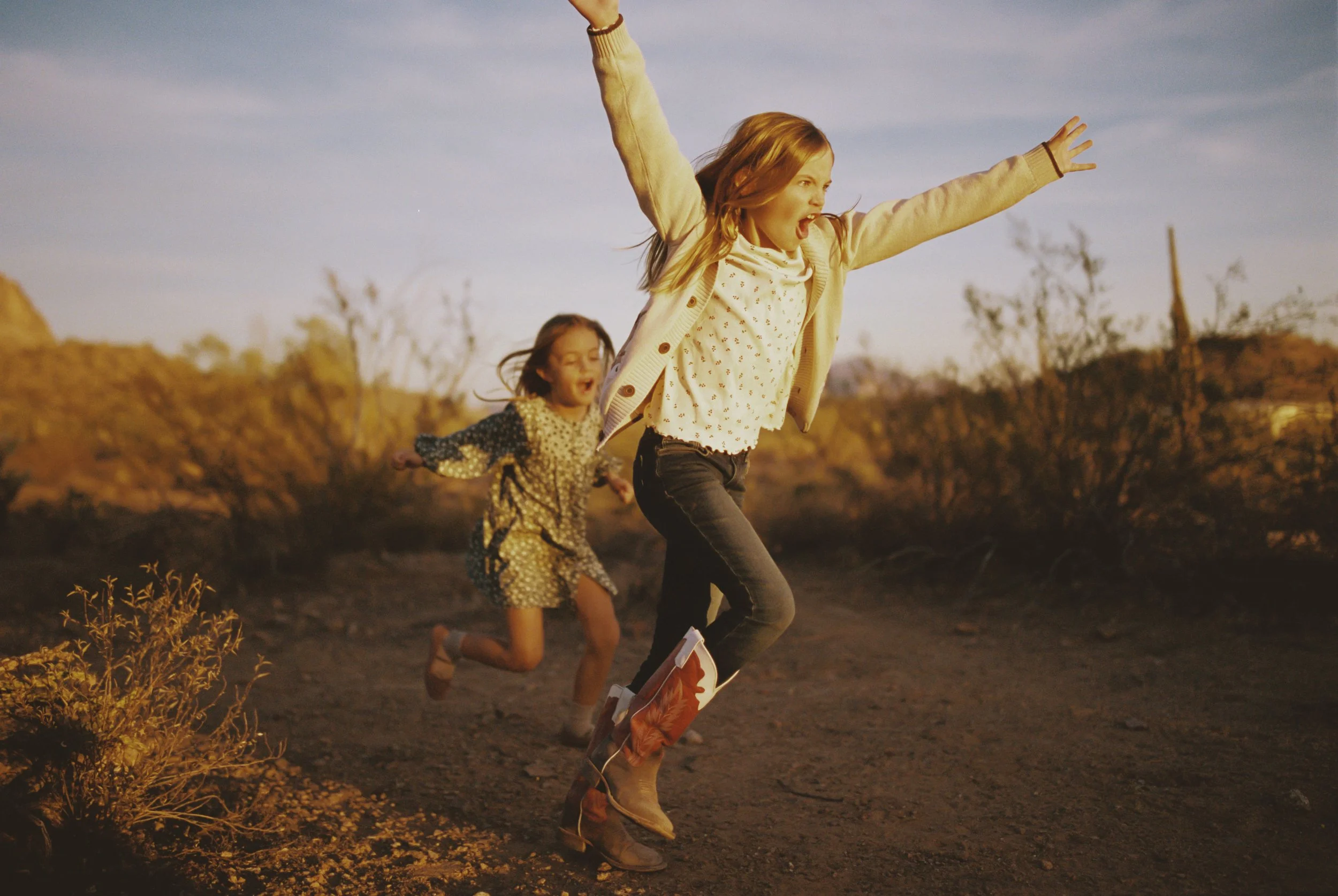 Two young girls running outdoors in a desert landscape during sunset, with one girl leading and the other following, both with joyful expressions.