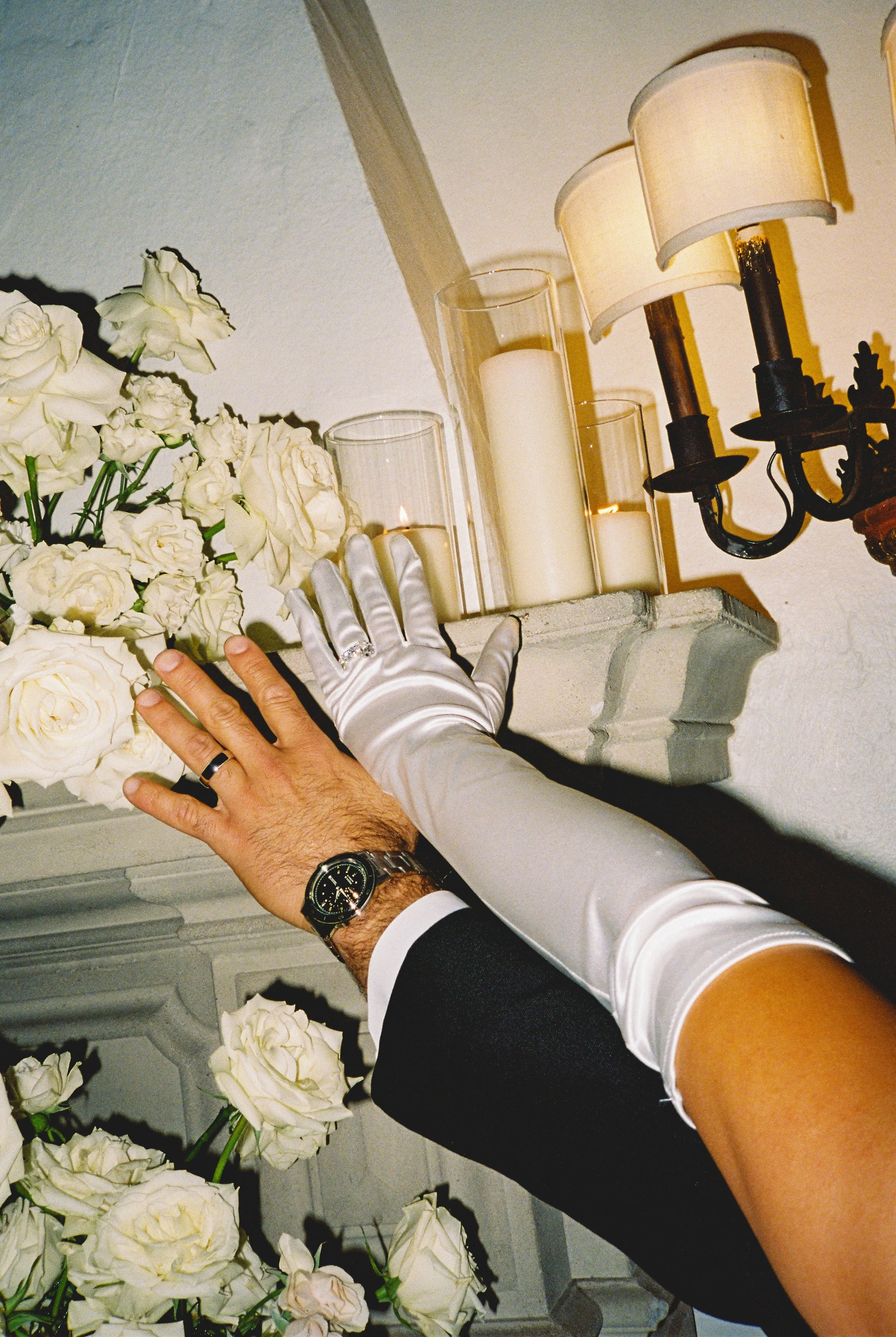 A couple's hands, one wearing a wedding band and a black wristwatch, touch a white floral arrangement with candles and lamps in the background.