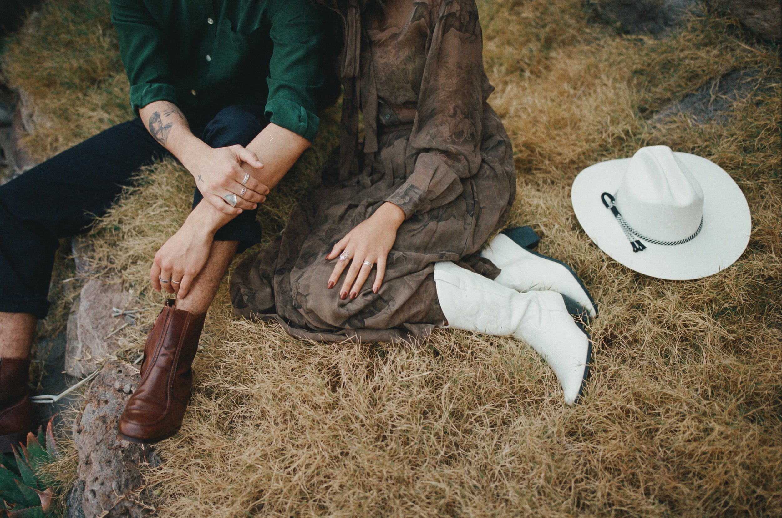 Close-up of two people sitting on dry grass. One person is wearing brown boots, black pants, and a green shirt. The other person is wearing a brown patterned dress and white boots, with a white cowboy hat nearby.