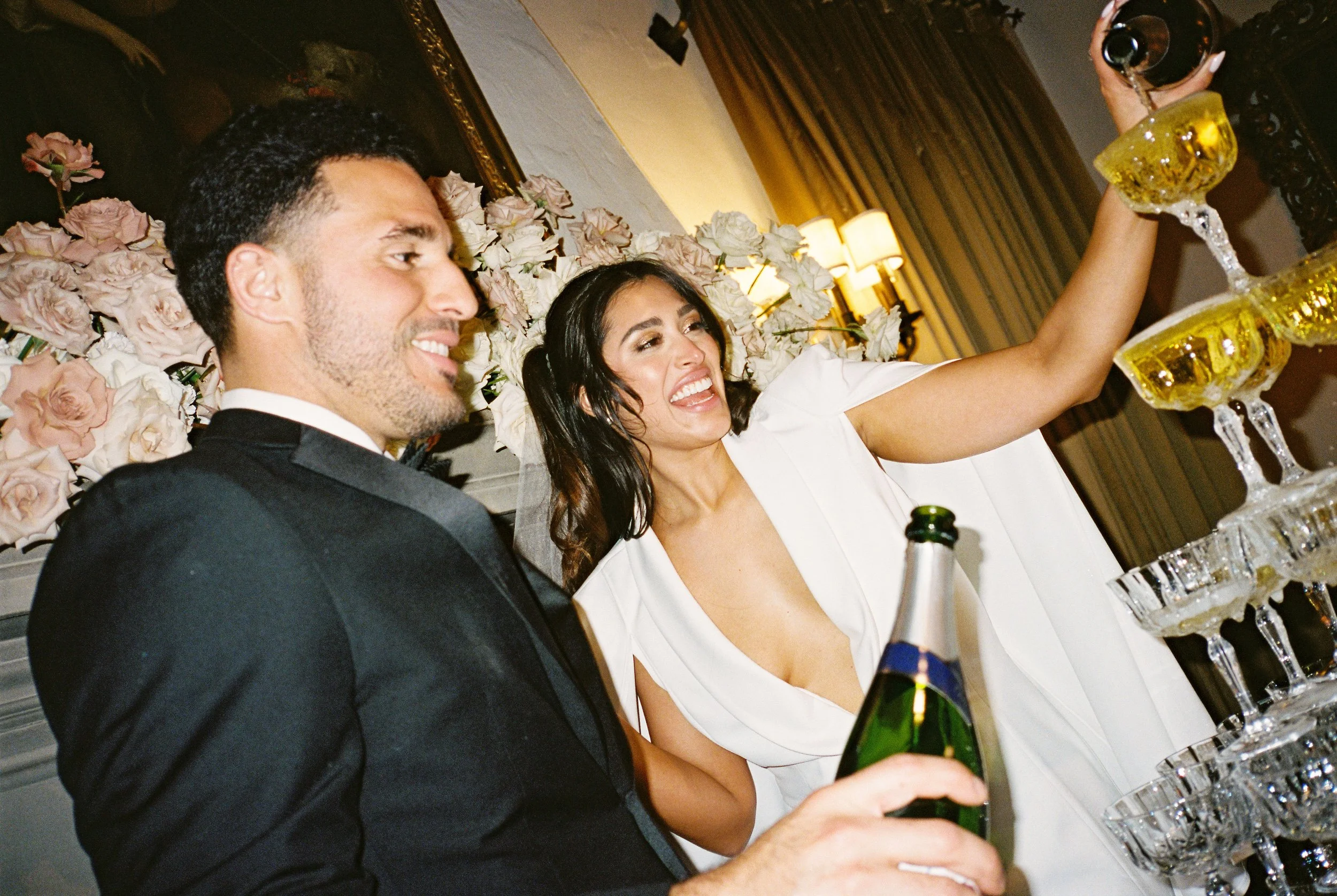 A couple in wedding attire celebrating with champagne and a tower of glasses, with a floral backdrop in a decorated indoor setting.