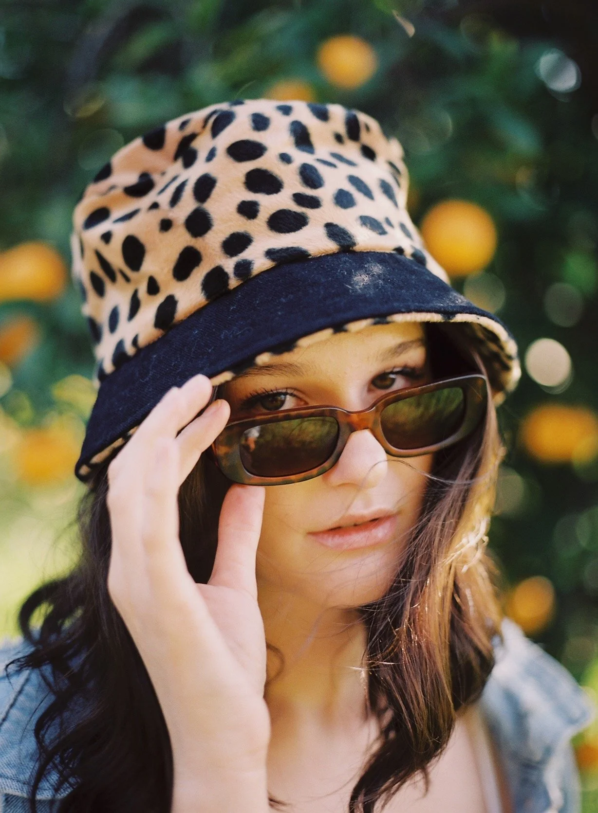 A woman with brown hair wearing leopard print bucket hat and sunglasses, holding the sunglasses with one hand, with an orange tree in the background.