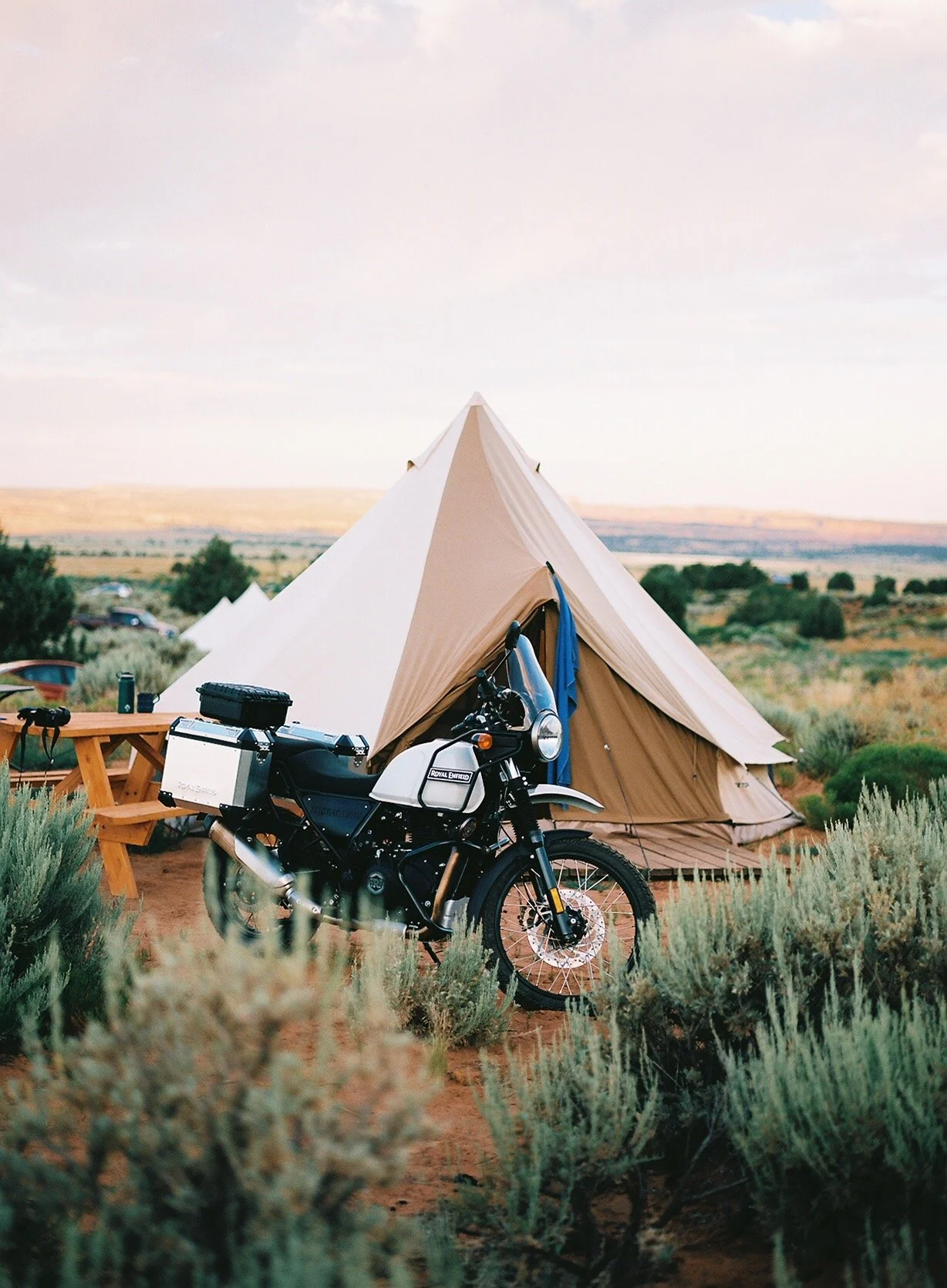 A black and white motorcycle parked in front of a beige canvas tent in a desert landscape with shrubs and a distant horizon.
