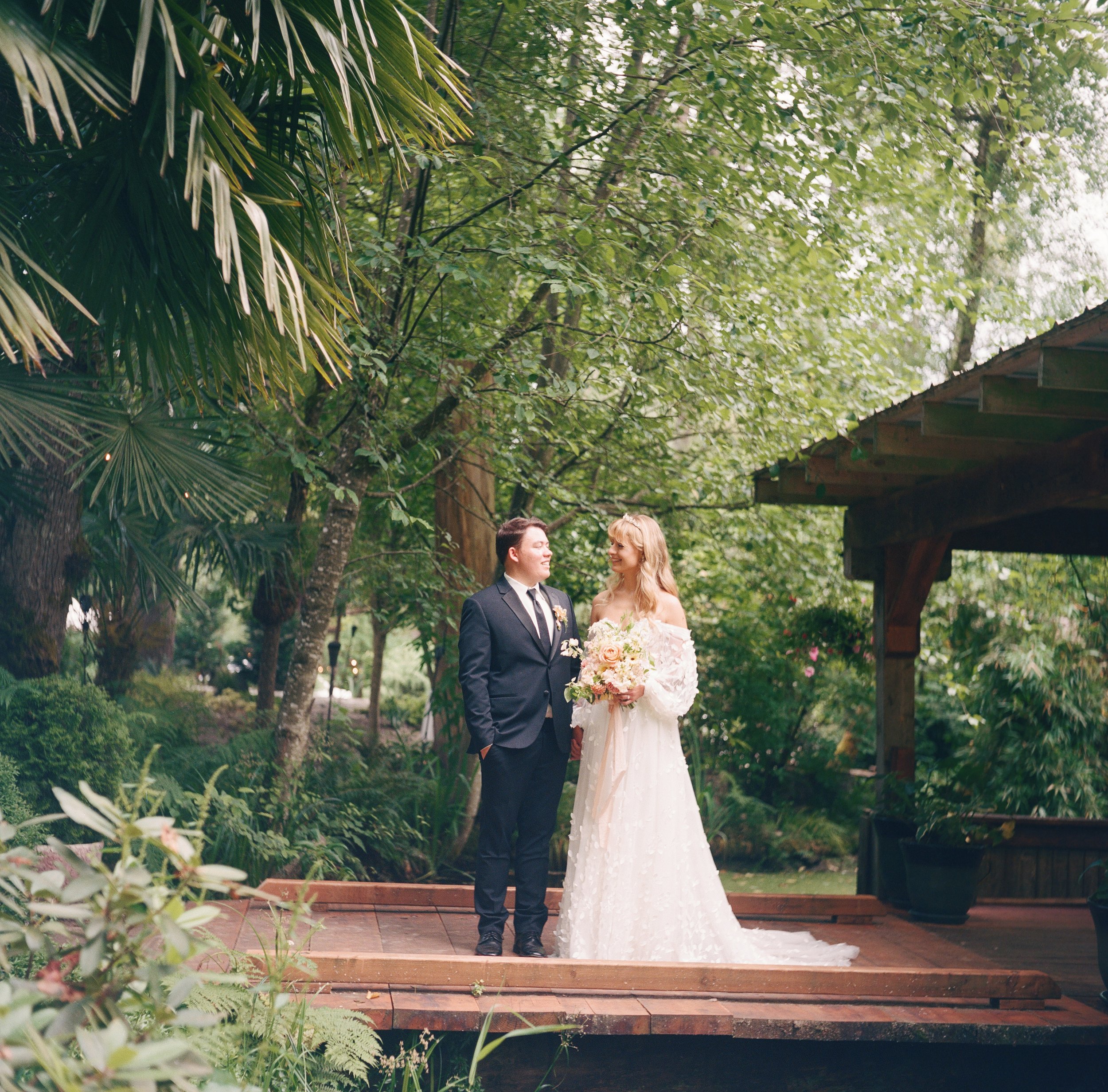 A bride and groom standing on a wooden platform in a lush, green garden. The bride is holding a bouquet and wearing a white wedding dress, while the groom is in a dark suit with a tie, both smiling at each other.