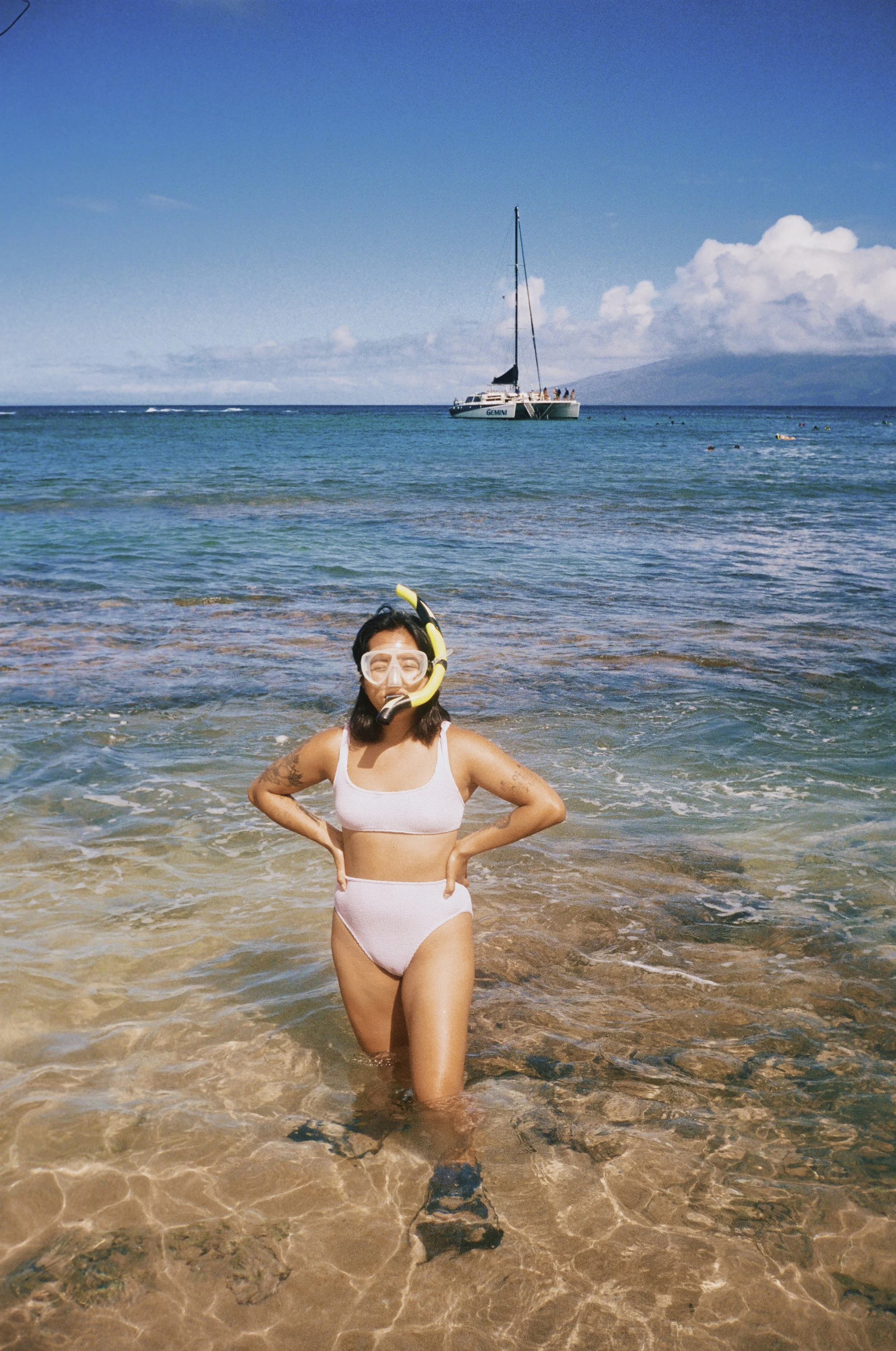 A woman in a white swimsuit with a snorkel and mask standing in shallow clear water on a beach, with a sailboat in the distance against a blue sky with clouds.