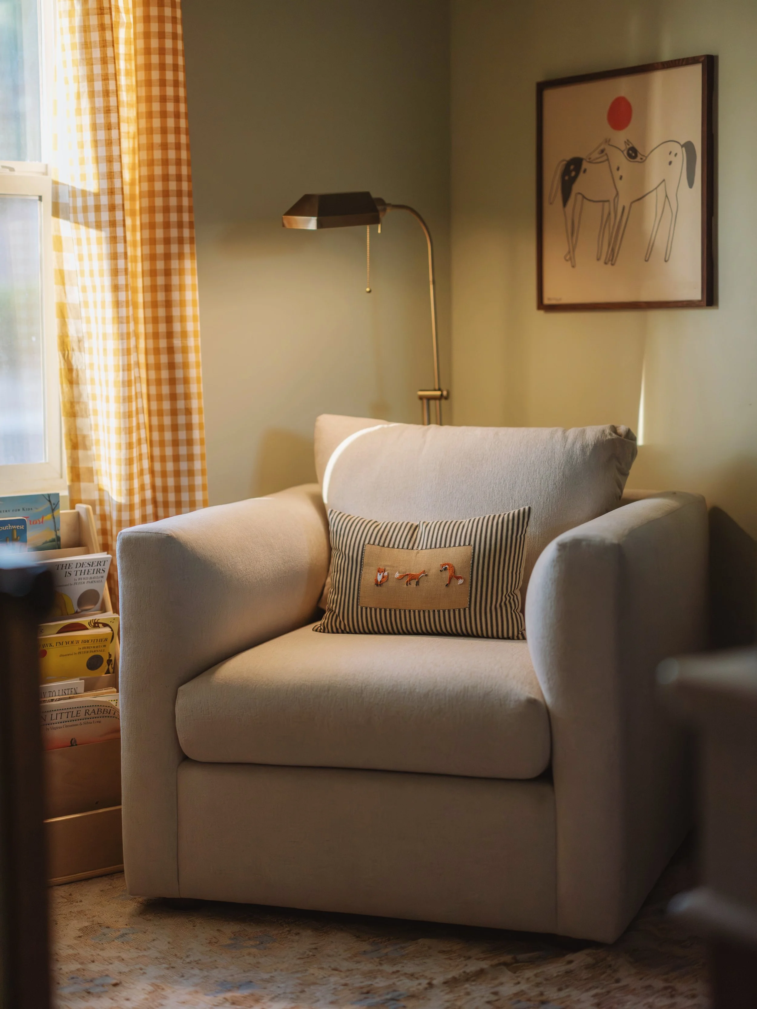 Cozy living room corner with a beige armchair, embroidered pillow, standing lamp, framed horse artwork, and a bookshelf with books near a window with yellow checkered curtains.