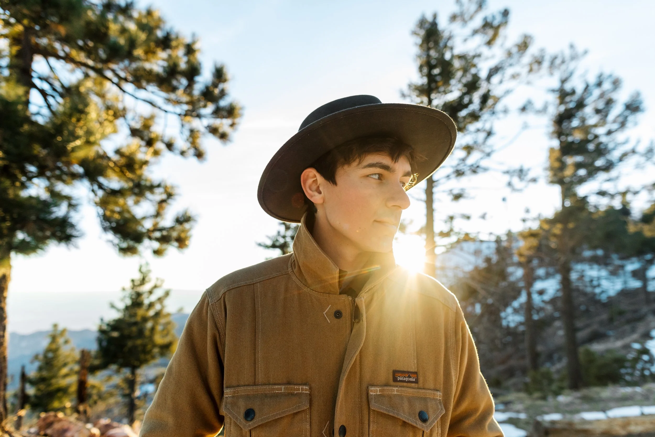 A young man wearing a wide-brimmed hat and a tan outdoor jacket standing in a forested area during sunset, with sunlight filtering through the trees in the background.