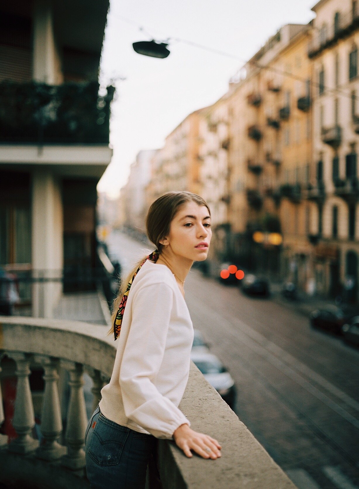 A young woman with brown hair tied back in a colorful scarf, wearing a white jacket, leaning on a balcony railing overlooking a city street with old buildings, parked cars, and blurred traffic lights.