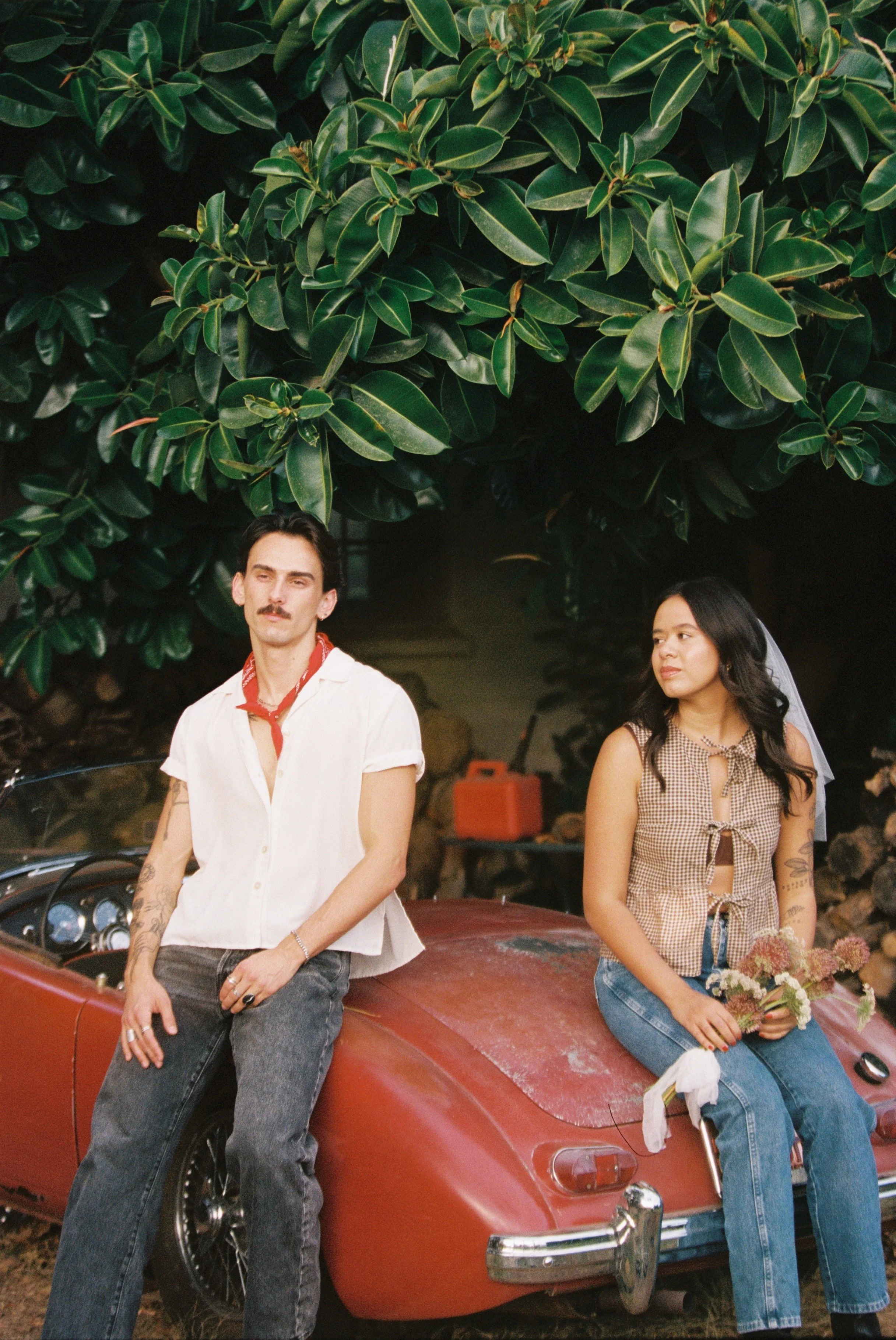 A man and woman sitting on a vintage red car outdoors under a large green leafy tree, with the woman holding a bouquet of flowers.