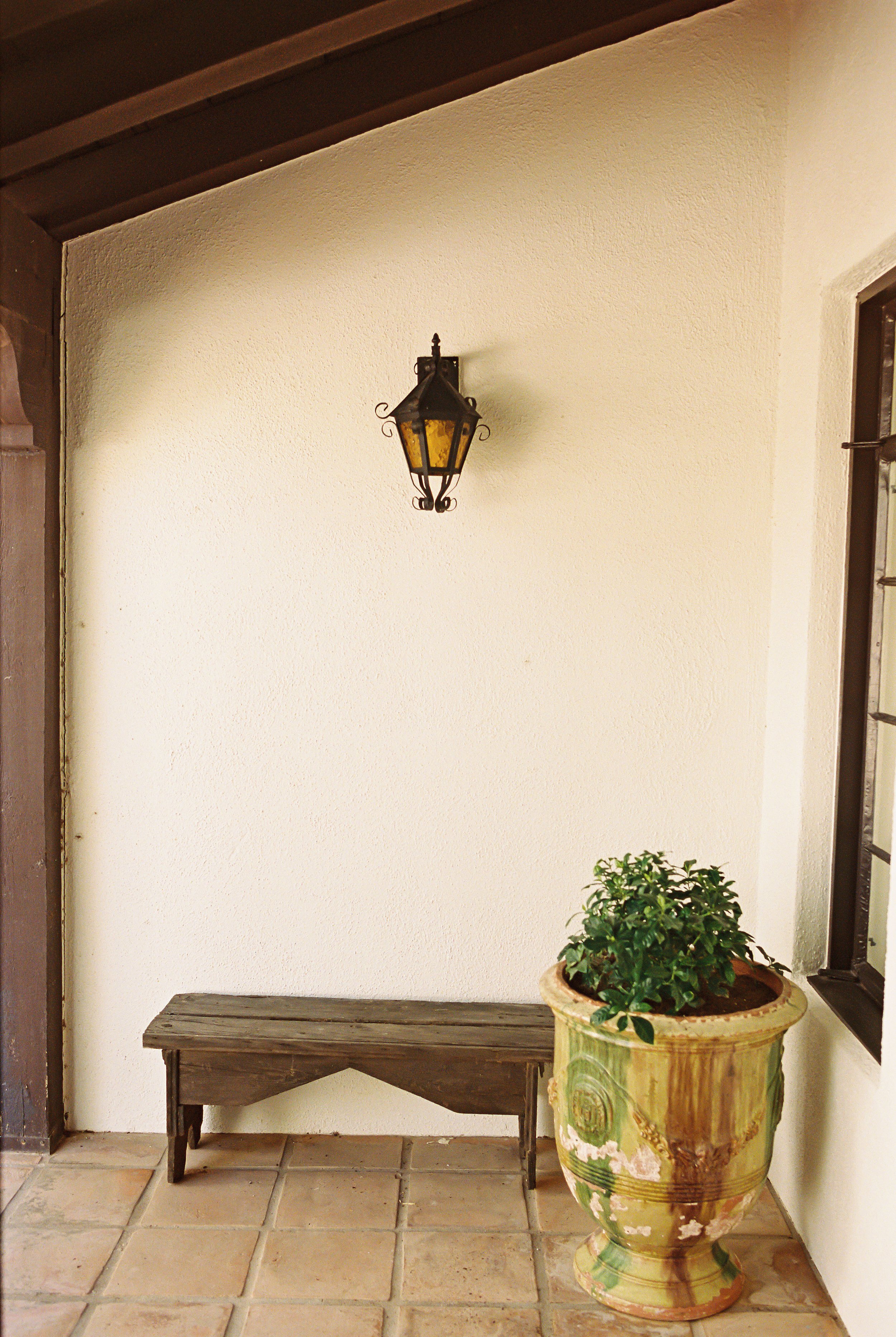 A small porch area with a rustic wooden bench, a decorative green ceramic planter with a leafy plant, a wall-mounted lantern-style light fixture, and a window with black trim.