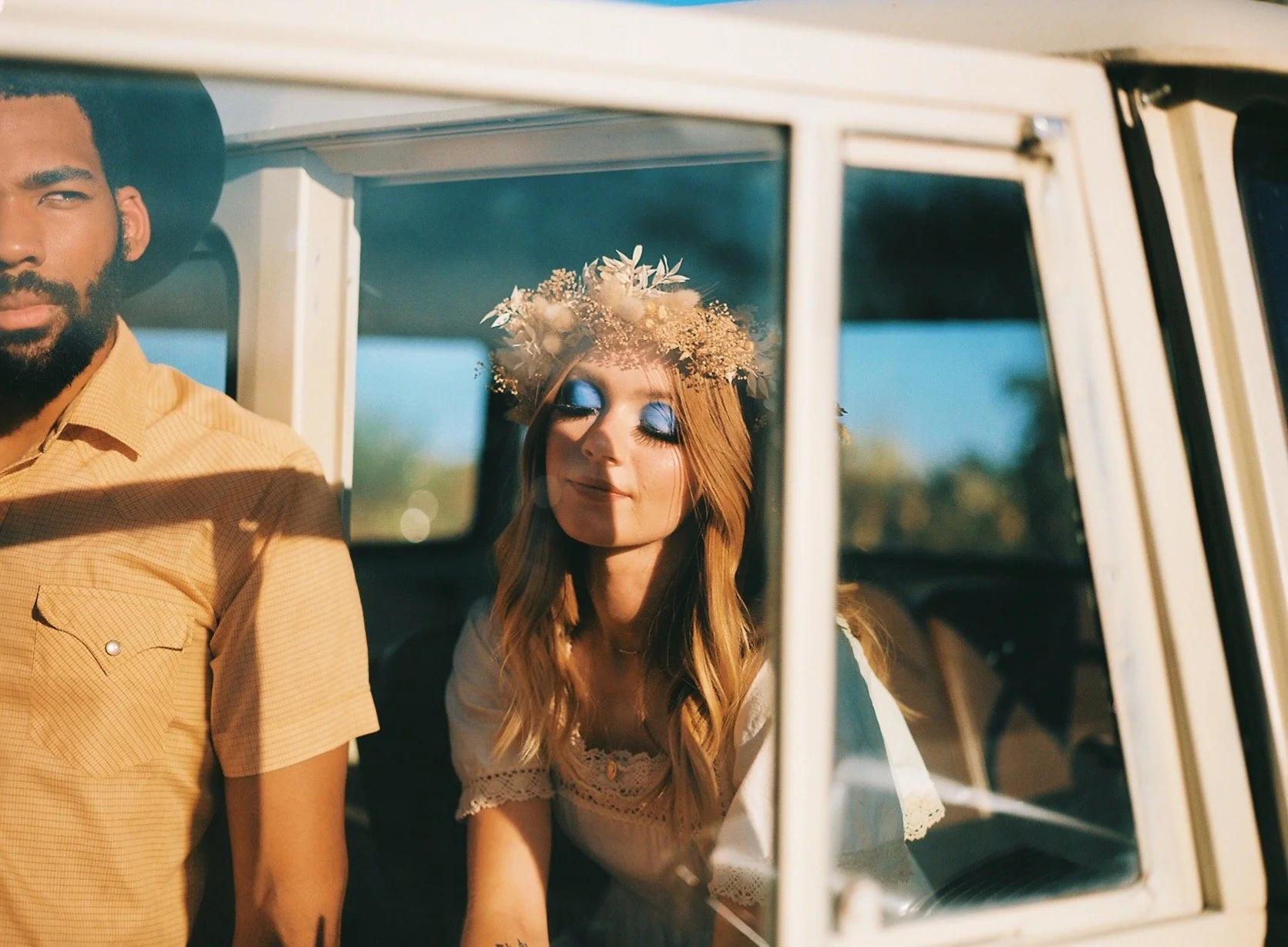 A young woman with red hair, blue eye makeup, and a floral headband is sitting inside a vehicle with her eyes closed, smiling softly. Part of a man wearing a yellow shirt and a black hat is visible on the left side of the image.