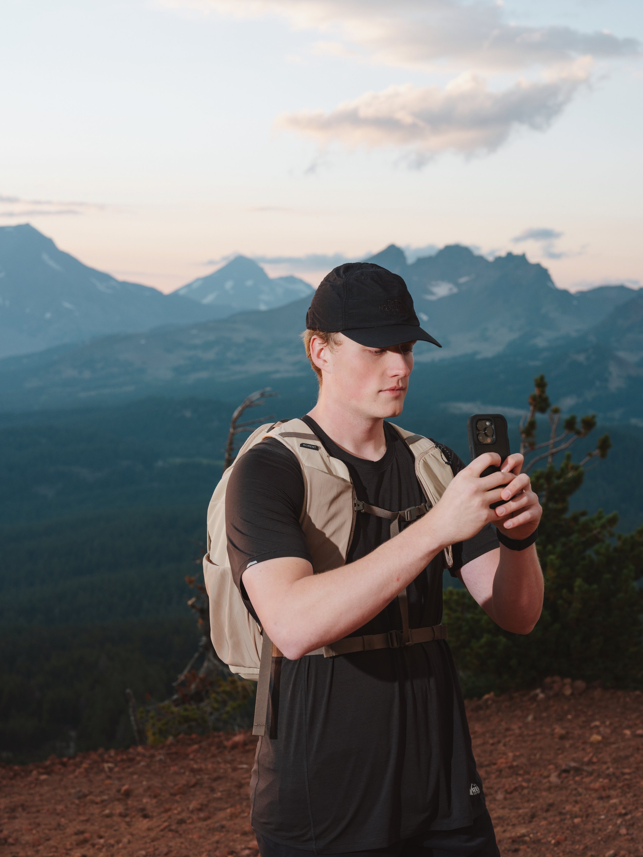 Young man in outdoor clothing using a smartphone on mountain trail with mountain range and cloudy sky in background.