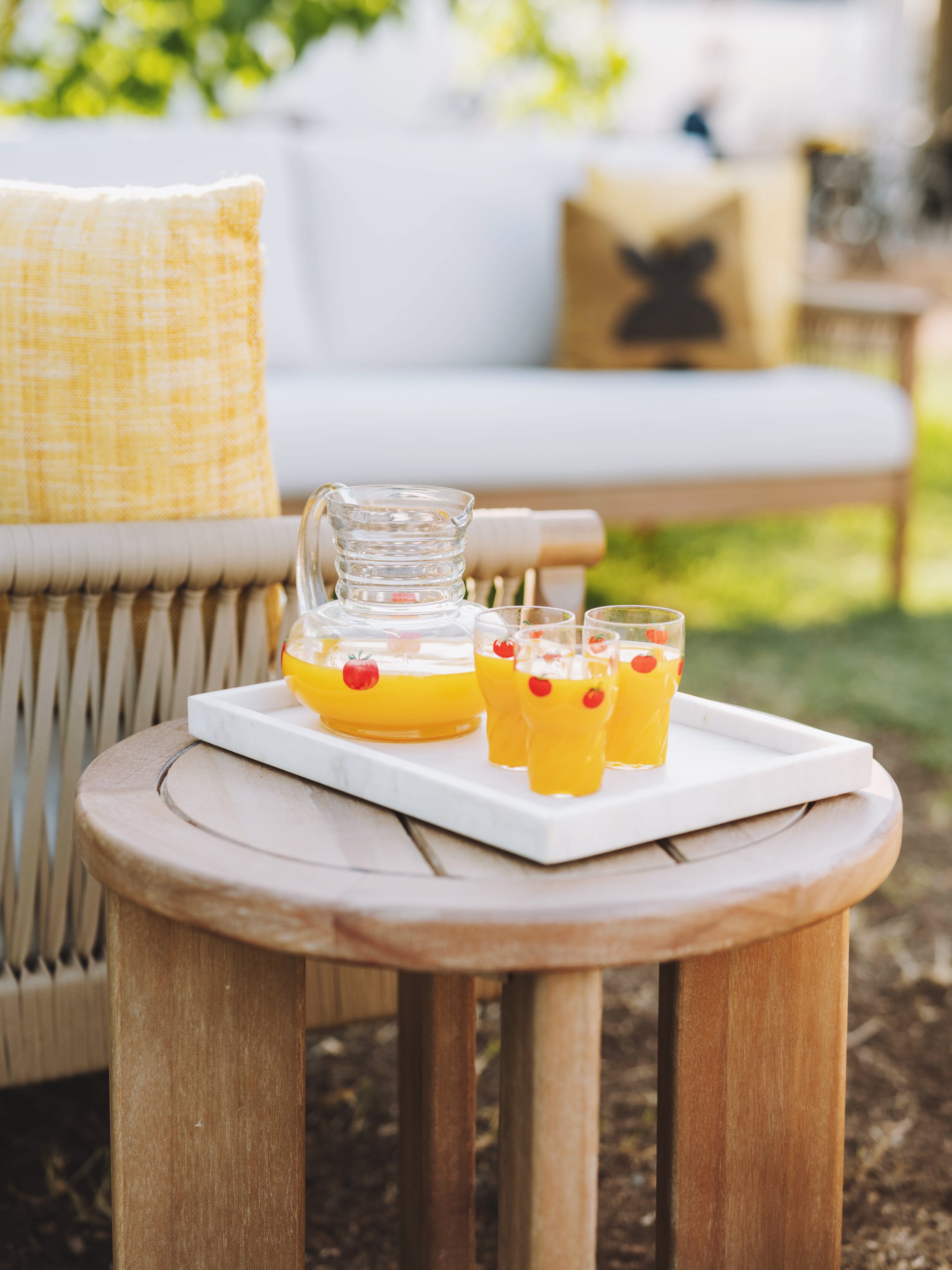 Glass pitcher and four glasses of orange juice with cherry decorations on a white tray on a round wooden side table outdoors, with a wicker sofa and yellow pillows in the background.