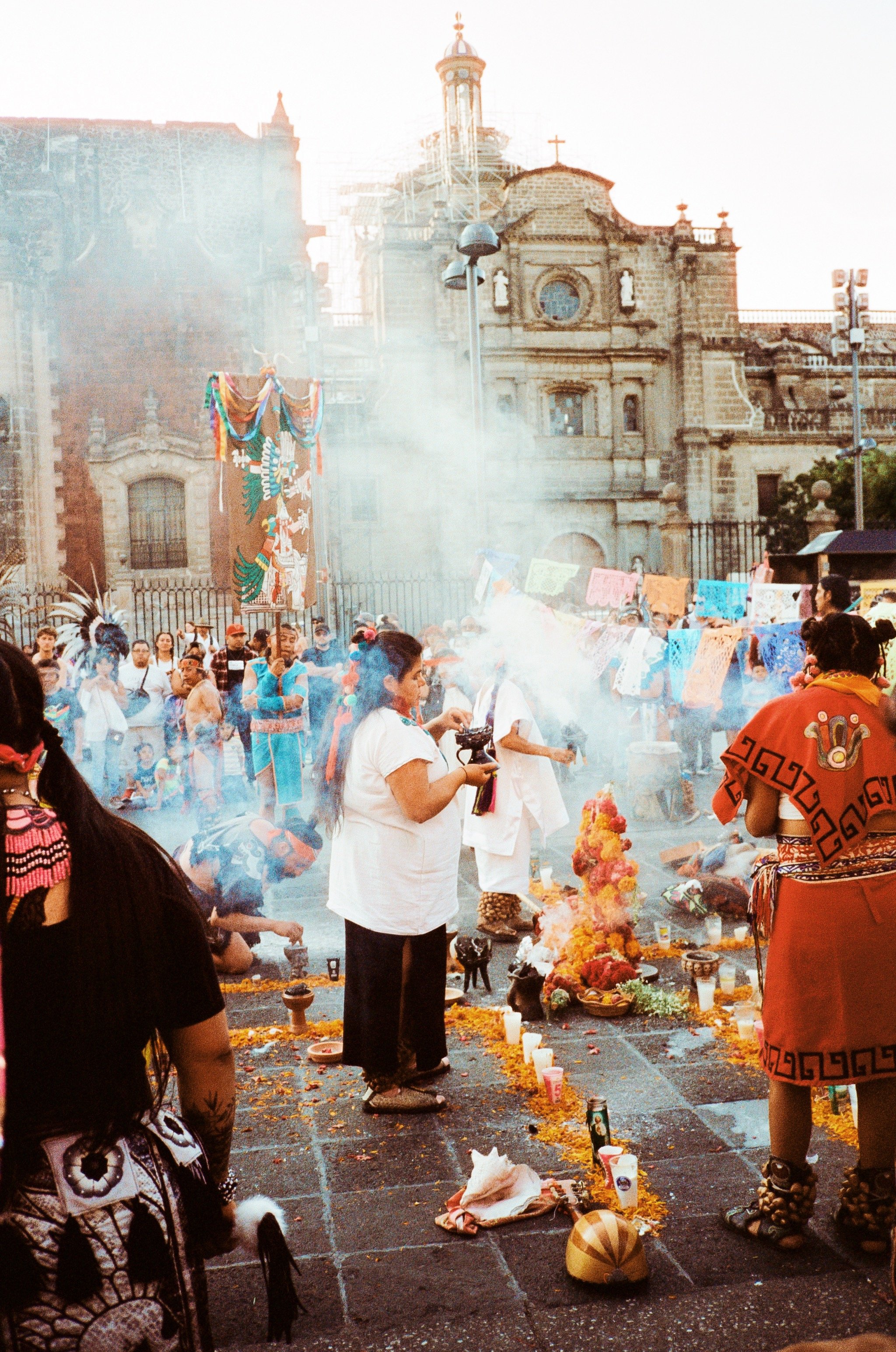 A group of people participating in a traditional Mexican celebration, including offerings, incense, and colorful decorations in front of a historic church with scaffolding.