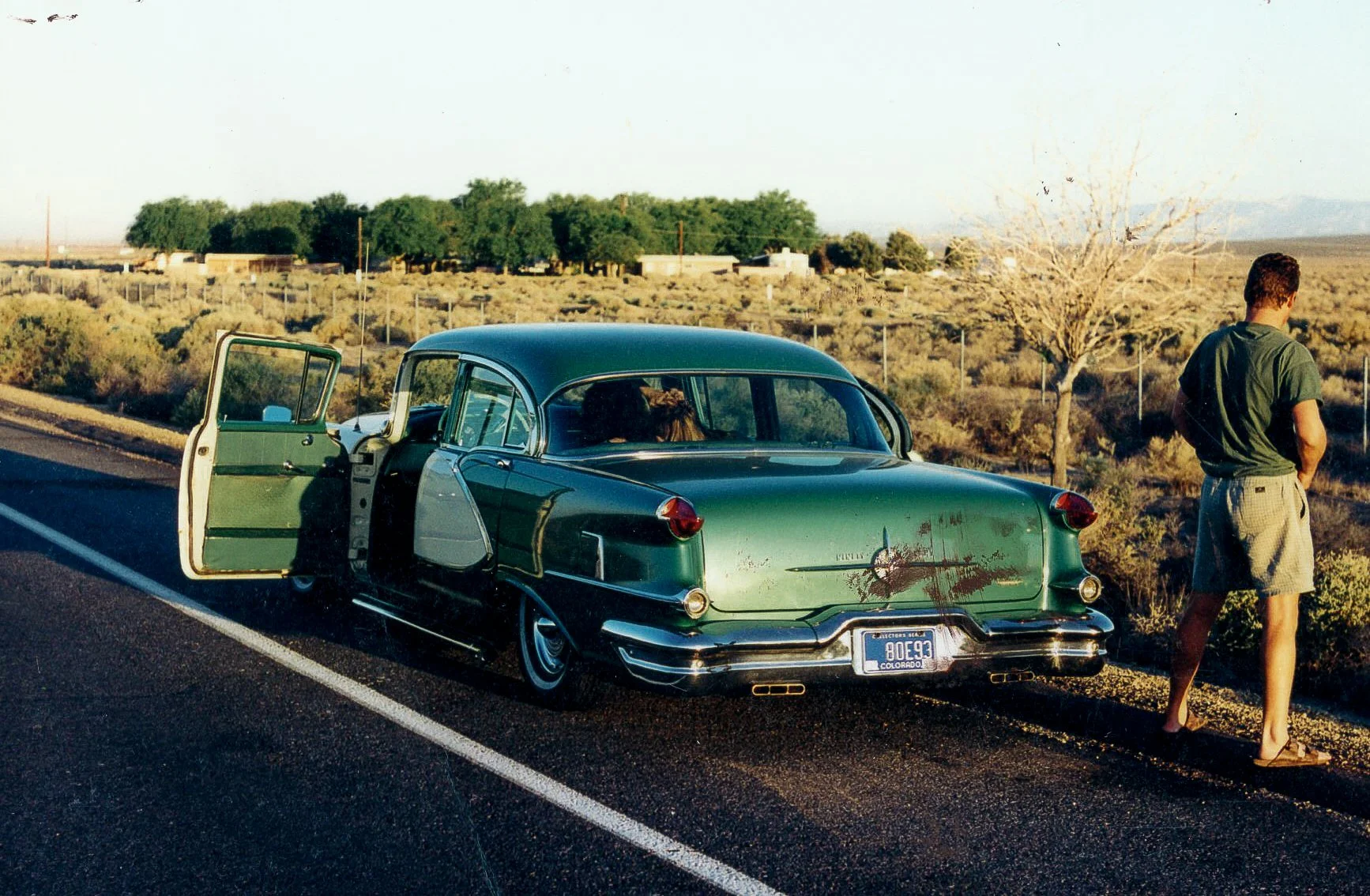 A vintage green car with damage on the trunk, parked on the side of a road in a desert landscape, with a man standing nearby looking away.