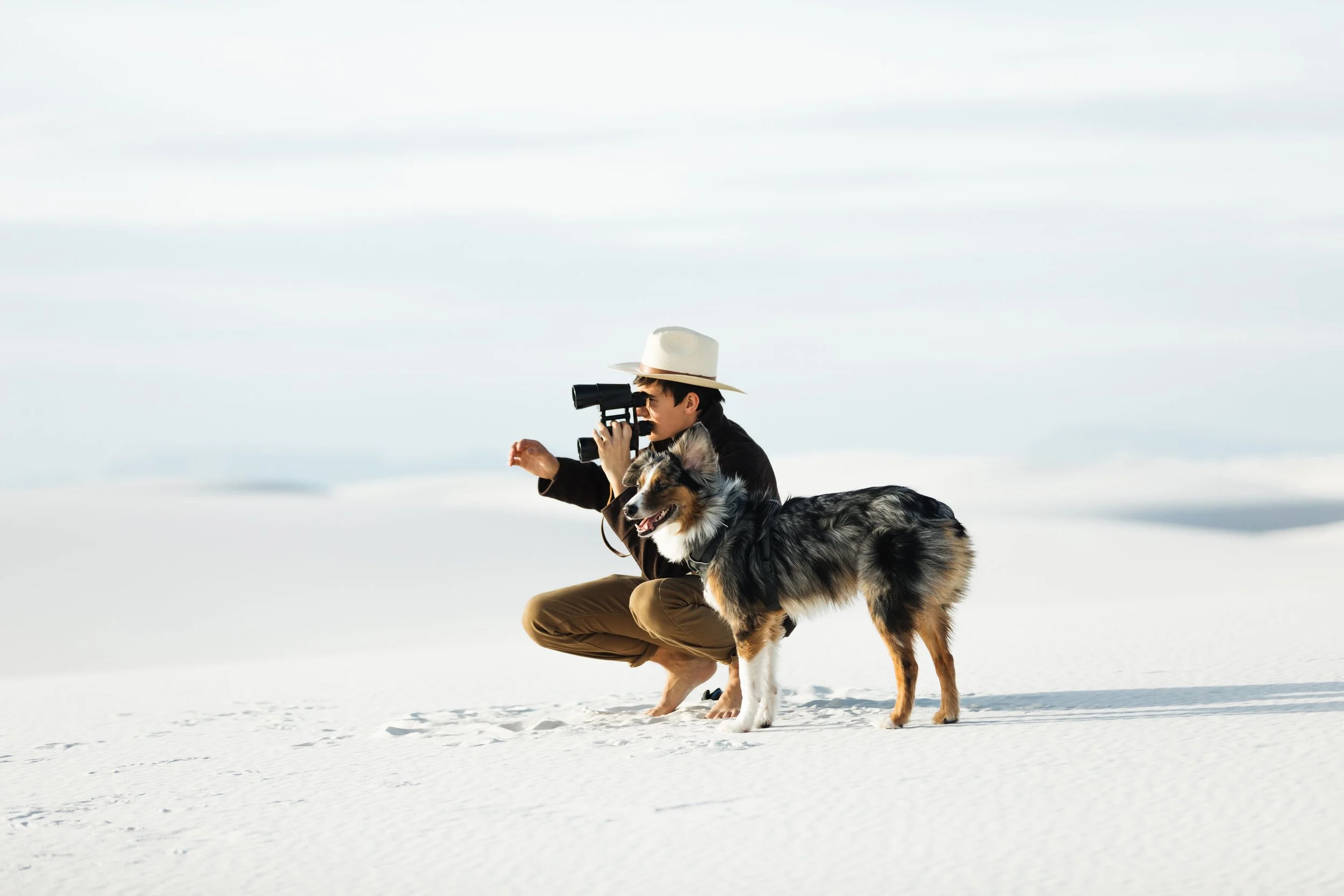 A person wearing a hat and sunglasses kneels in the snow, looking through binoculars, with a Border Collie standing next to them, in a snowy landscape.