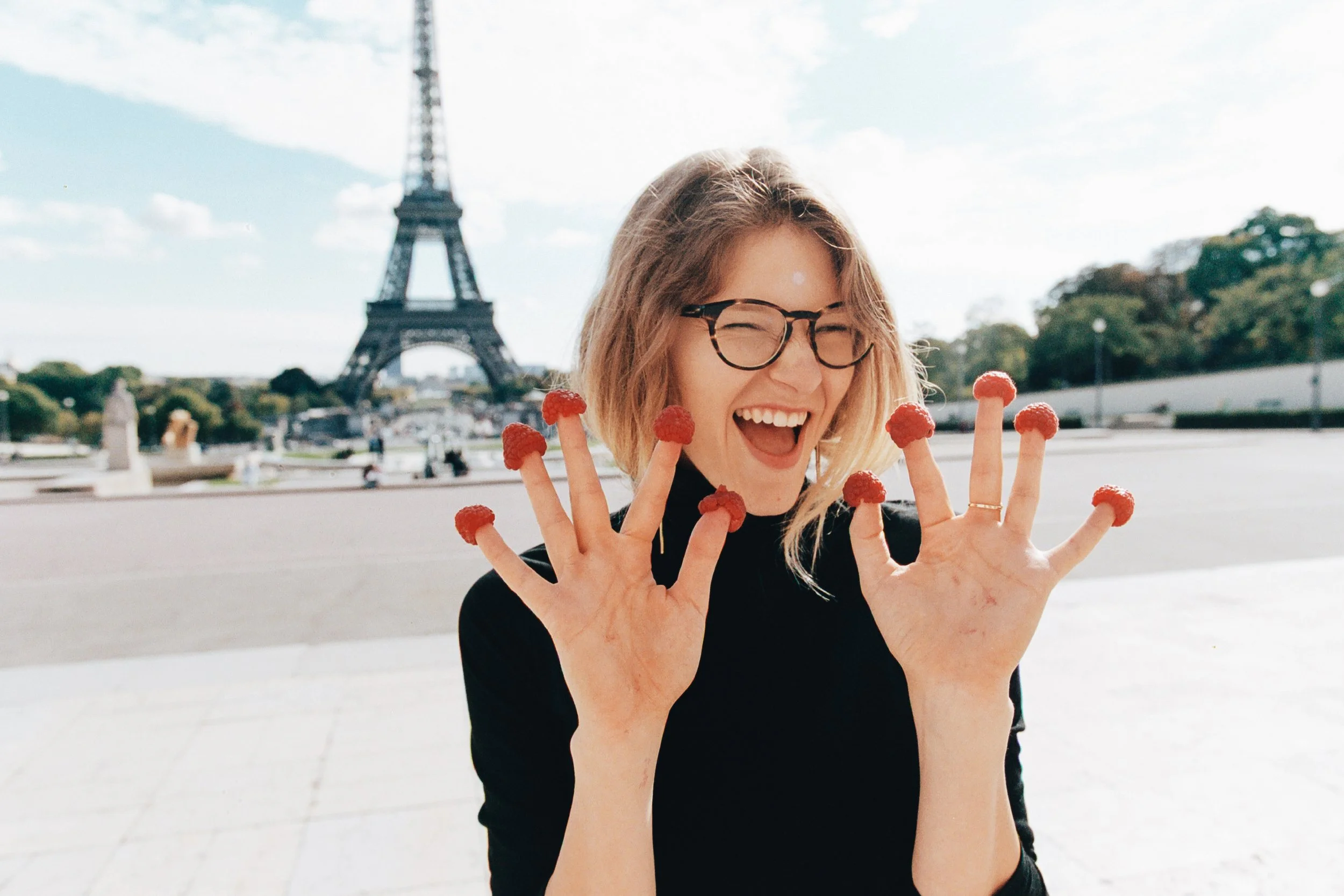 A woman with glasses and wavy blonde hair smiling with her eyes closed, holding up both hands with strawberries on her fingertips, with the Eiffel Tower in the background.