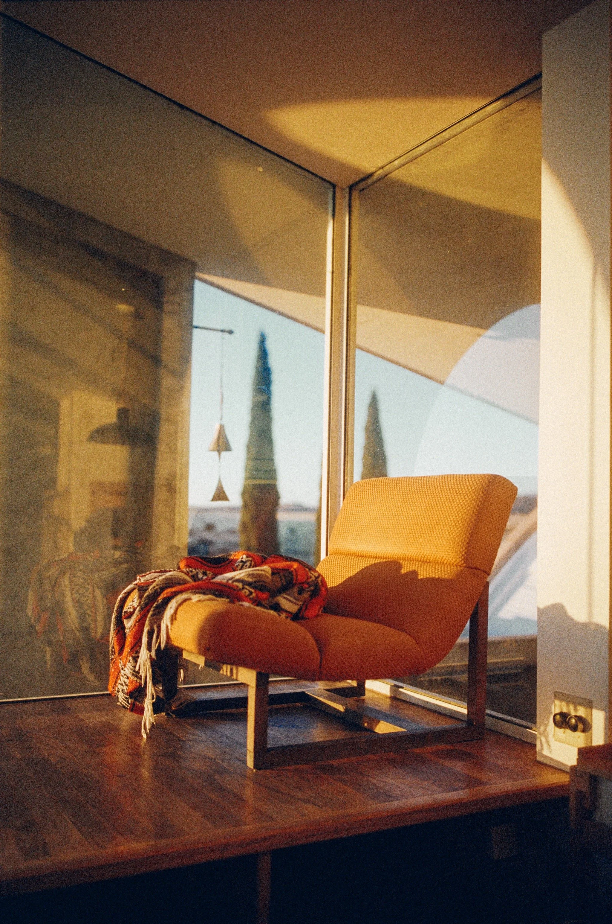 Sunlit room with orange lounge chair, colorful throw blanket, large glass window, and view of outside trees and blue sky.