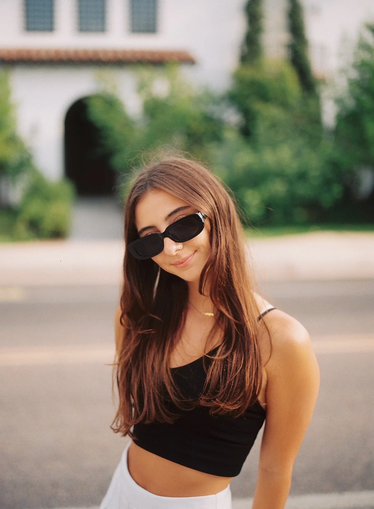 A young woman with long brown hair wearing black sunglasses, a black crop top, and white pants, smiling and standing outdoors with a blurred house and greenery in the background.