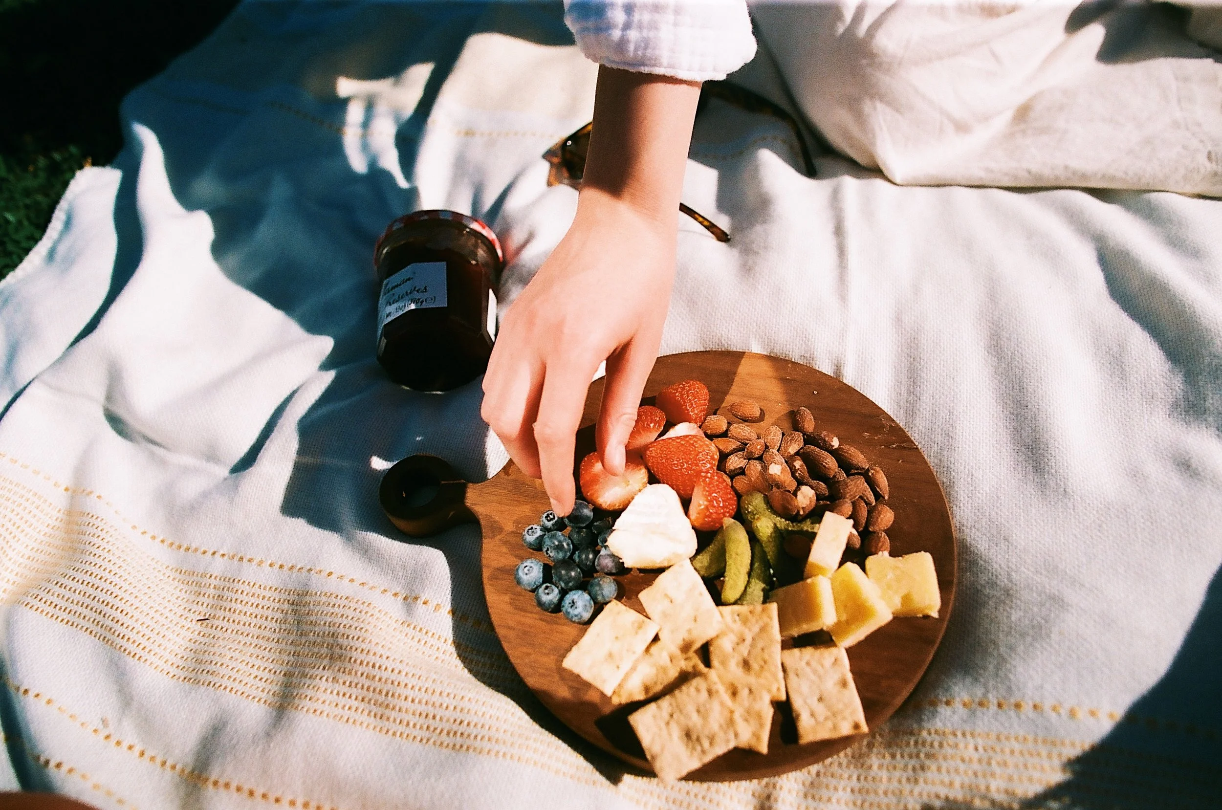 Person's hand reaching for strawberries on a wooden cheese board with grapes, almonds, and cheese pieces, placed on a white cloth, with a small jar and dark item nearby.