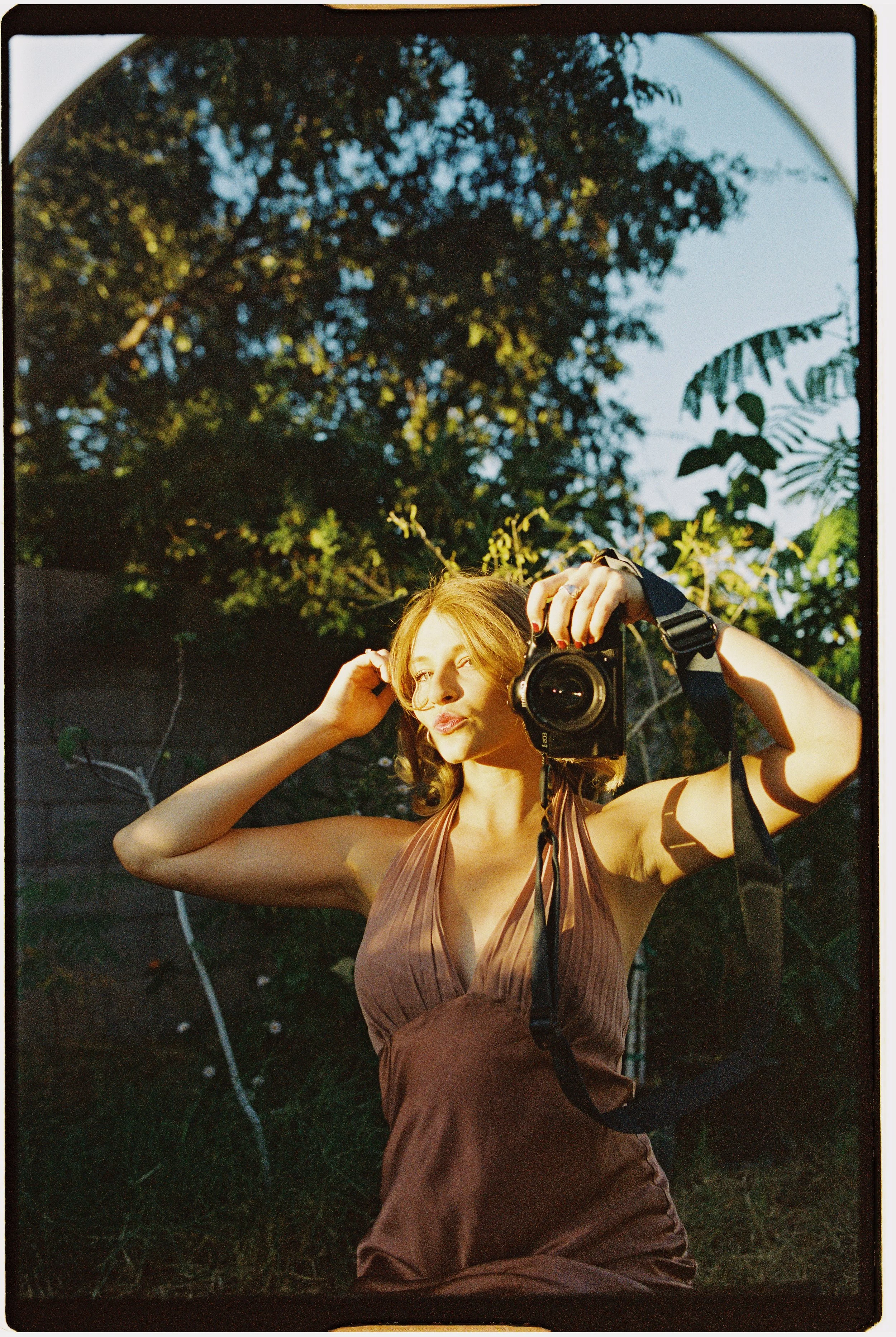 A young woman in a tan dress is outdoors, taking a selfie with a digital camera, with sunlight illuminating her face and greenery in the background.