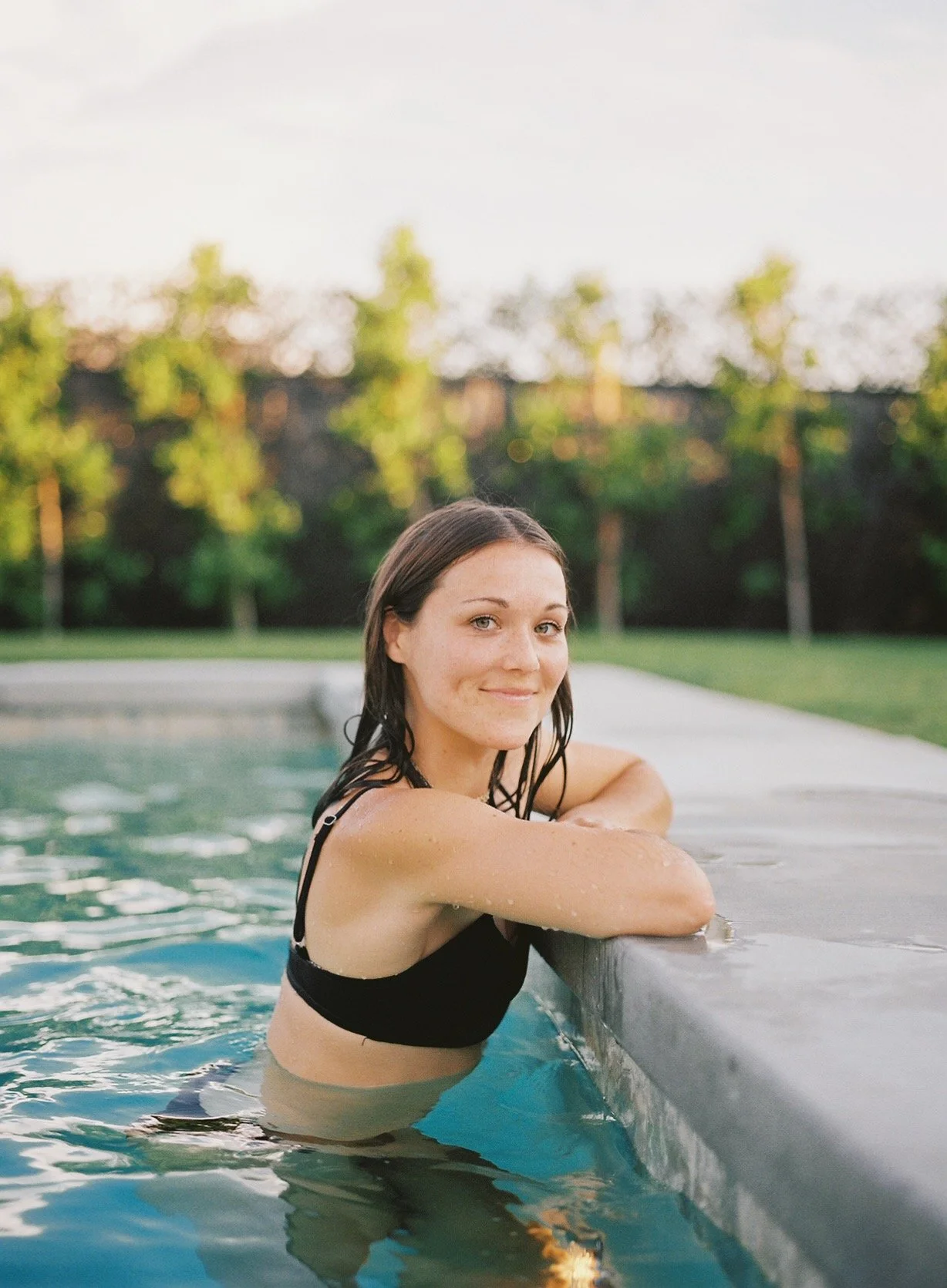 A woman with wet dark hair, wearing a black swimsuit, leaning on the edge of a swimming pool, smiling at the camera with trees and a cloudy sky in the background.