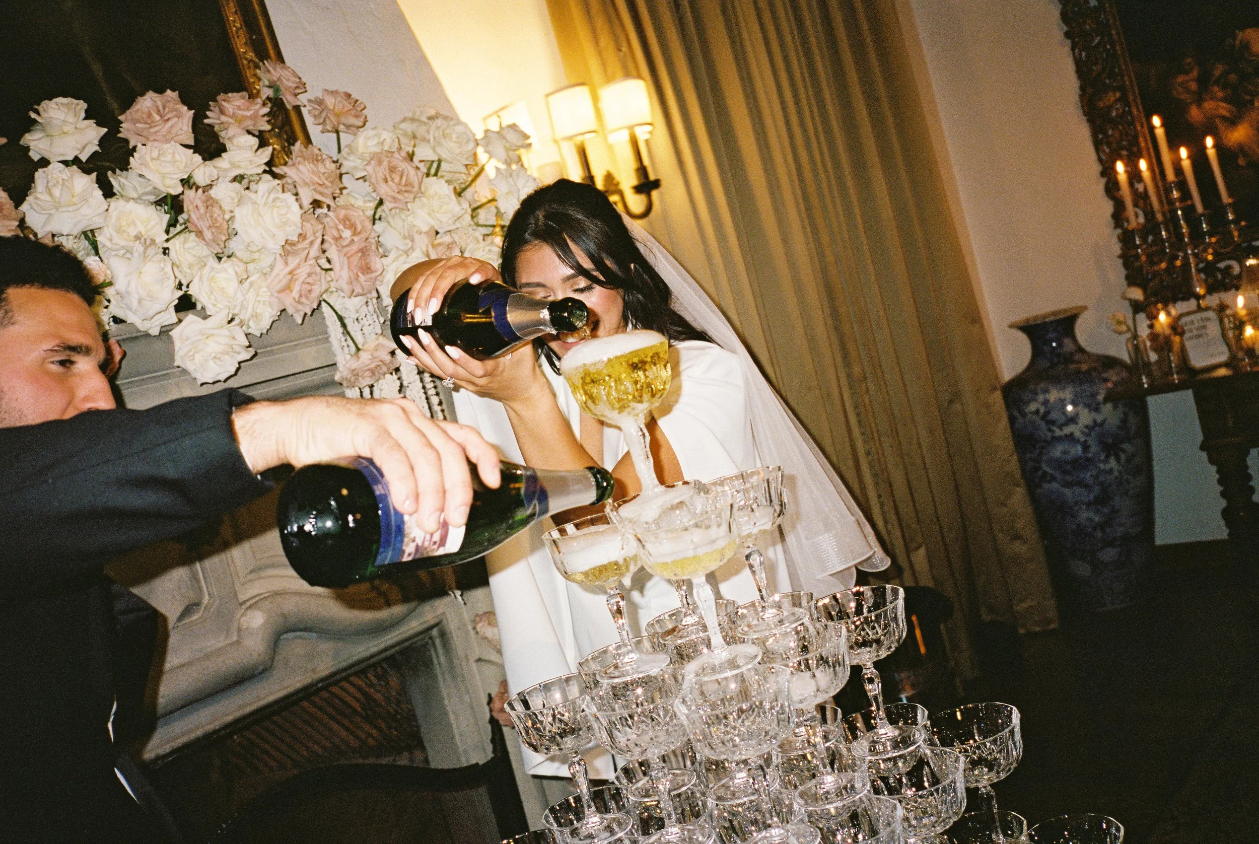A woman in a white dress and veil is pouring champagne into a tower of glass cups at a celebration event, with a man in black extending his arm to pour alongside her. The background features floral arrangements and elegant decor.