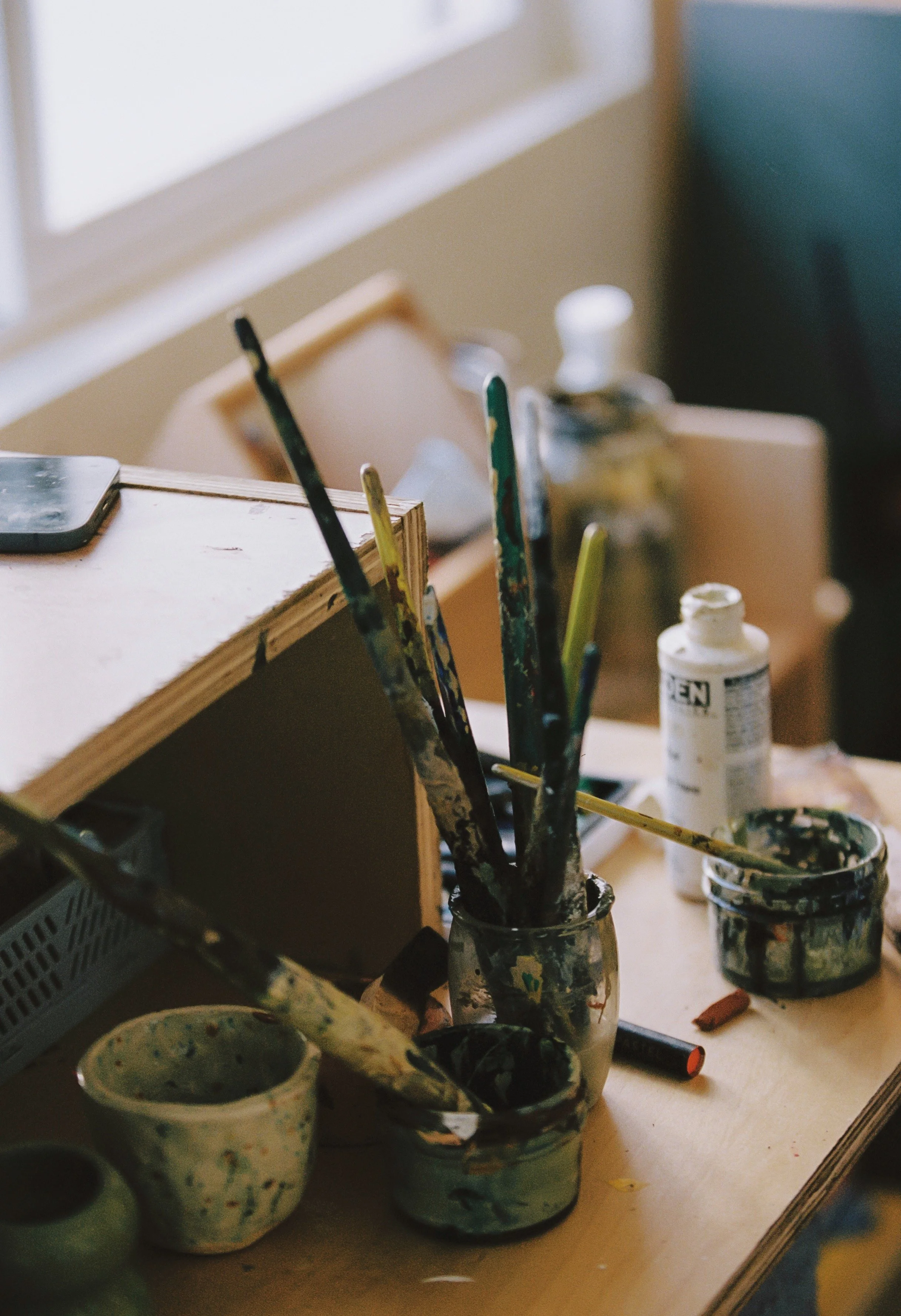 Art studio workspace with paintbrushes, jars, and paint bottles on a wooden table near a window.