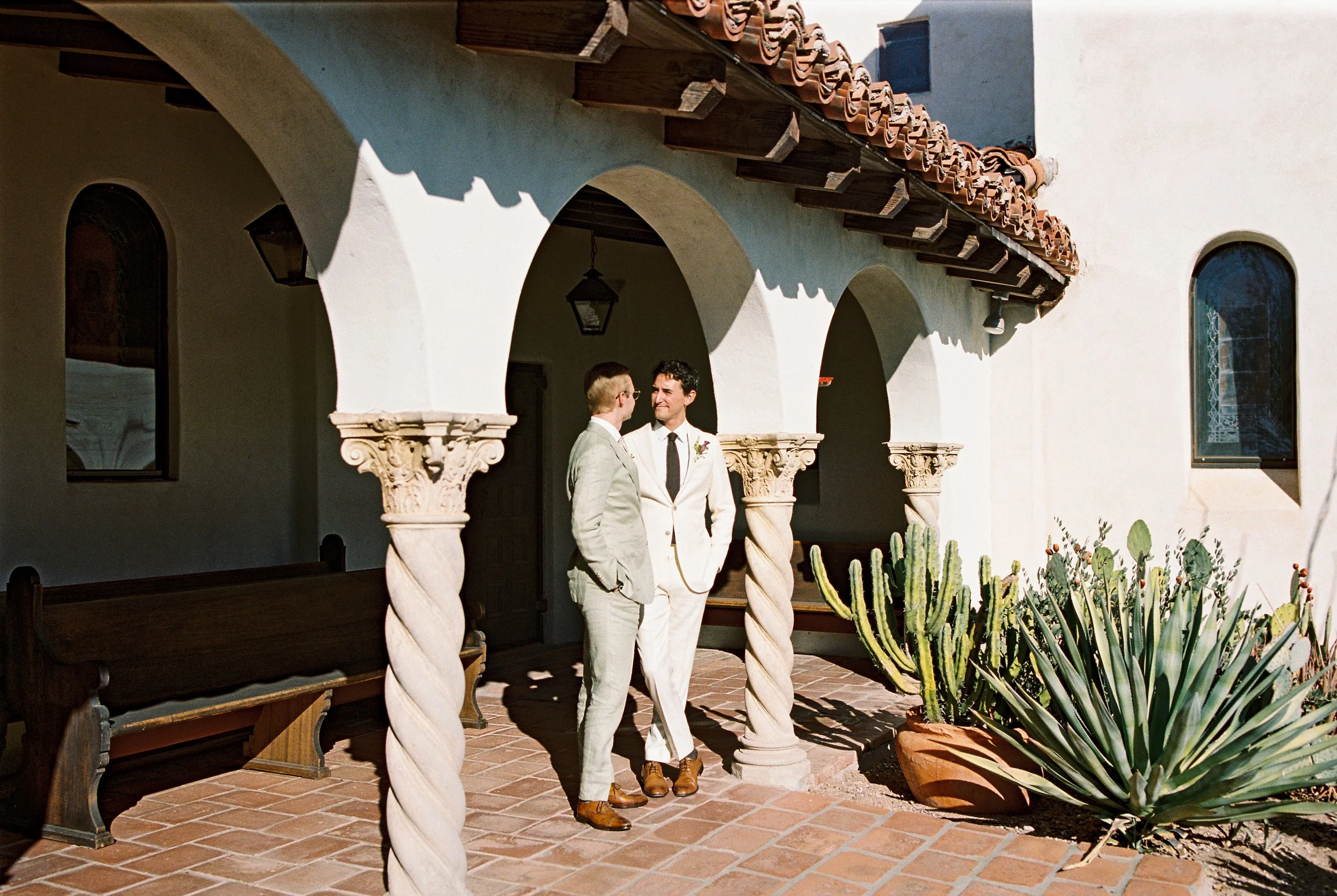 Two men in suits standing and talking under a covered Spanish-style porch with clay tile roof, stone columns, and desert plants nearby.