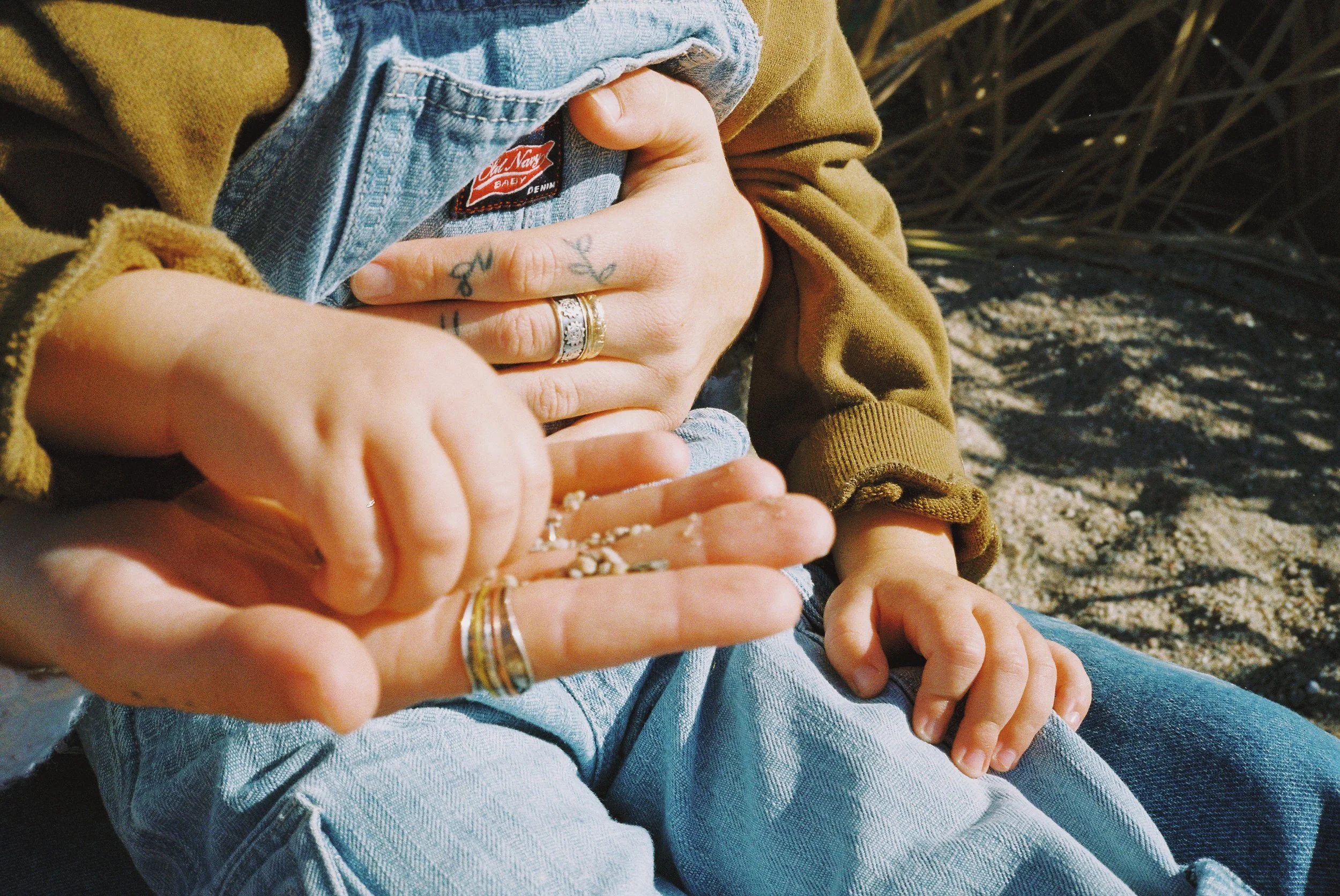 Close-up of a person holding a child on their lap, with both hands placed on the child's chest, while the child's hand is touching sand. The person is wearing rings, and the child is wearing a tan jacket and denim jeans. Sand and plants are visible i