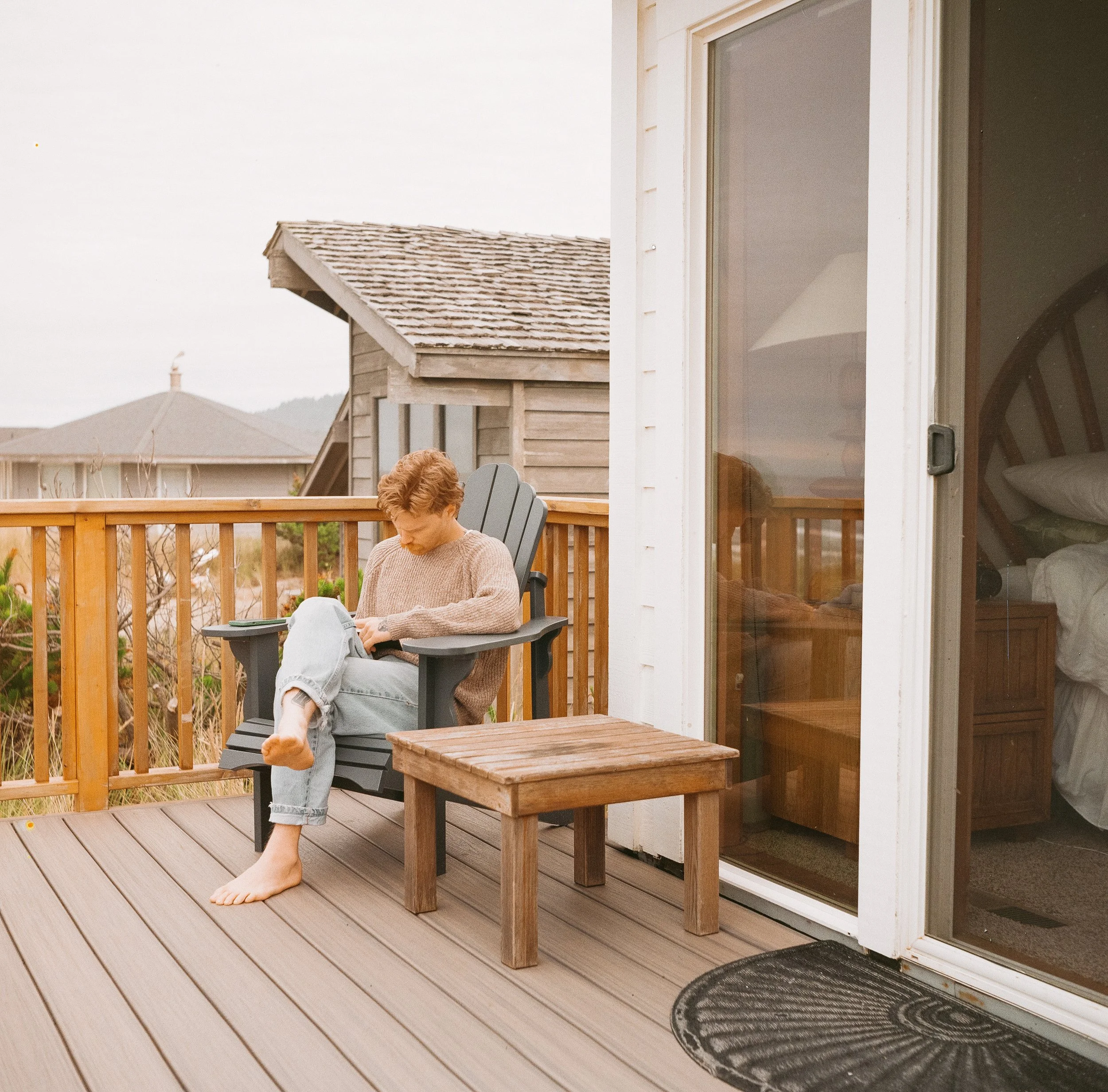 Person sitting on a gray wooden Adirondack chair on a wooden deck, reading a book during daytime. The deck is outside a house with sliding glass doors, revealing a bedroom interior. A small wooden table is beside the chair, and a black outdoor rug is