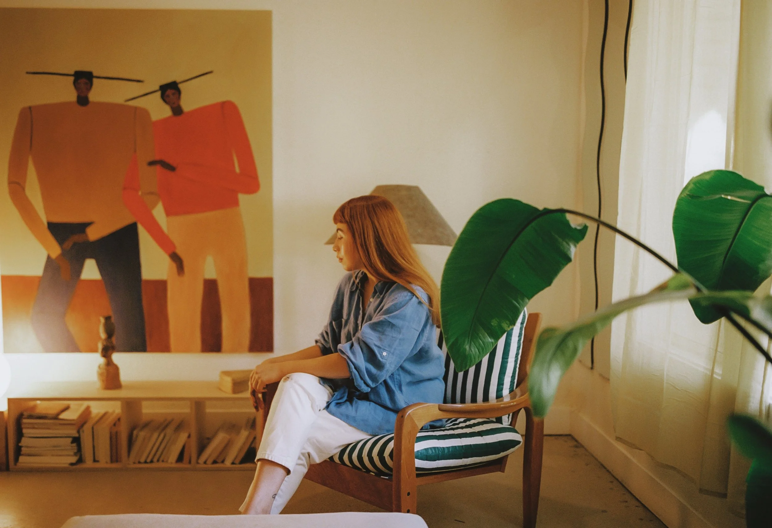 A woman with long red hair wearing a blue shirt and white pants sitting on a test green-and-white striped armchair in a living room, with a large leafy plant in the foreground and colorful artwork on the wall behind her.